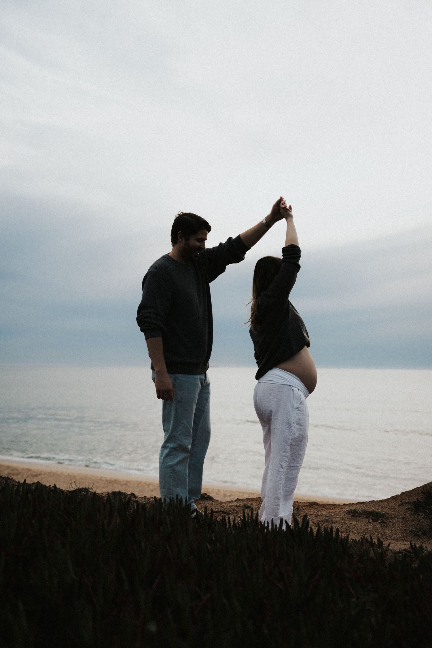 Pregnant couple dancing together on a coastal cliff overlooking the ocean on a cloudy day.