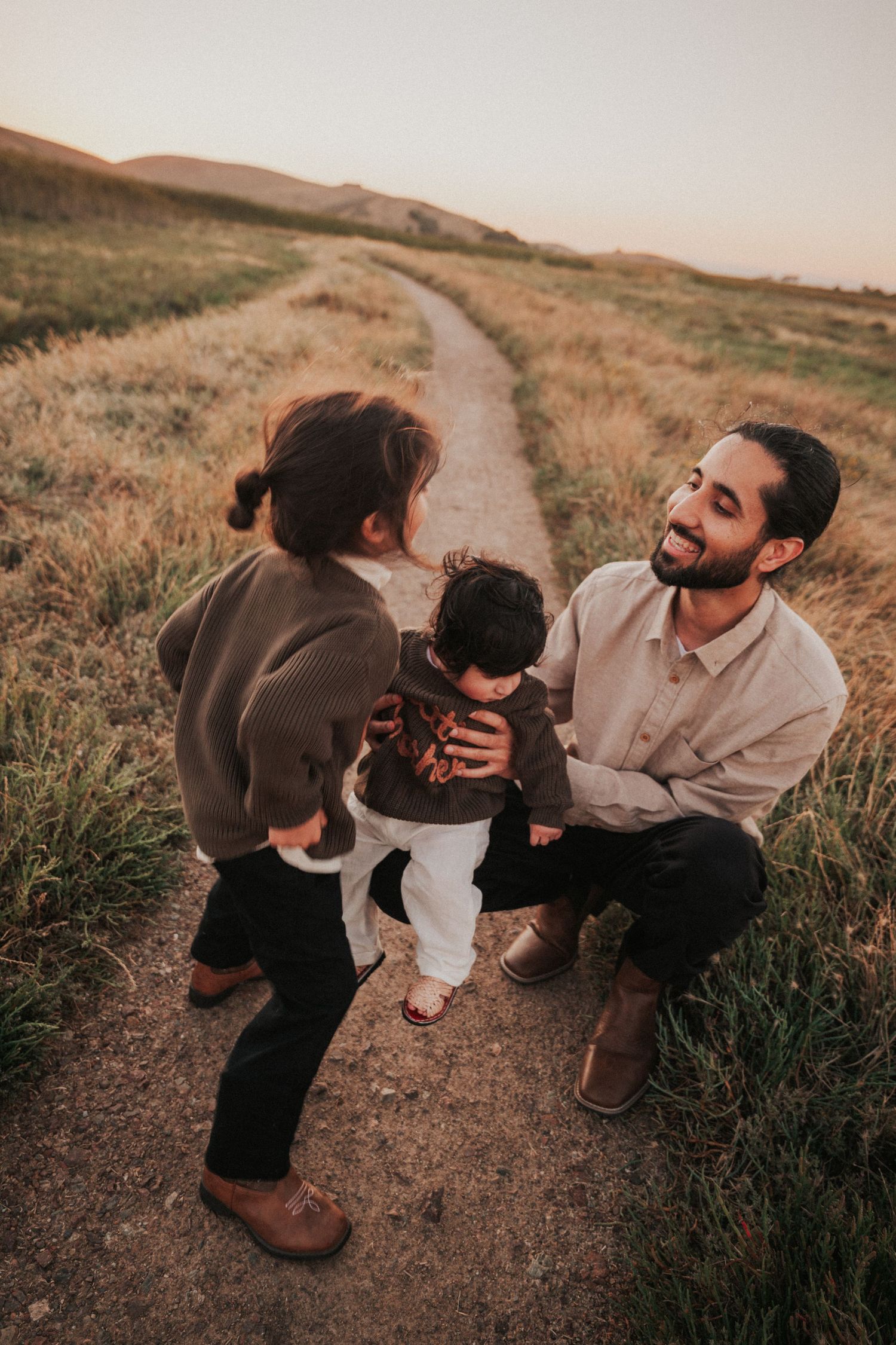 Smiling bearded father crouches on dirt path holding baby while toddler stands nearby at golden sunset.