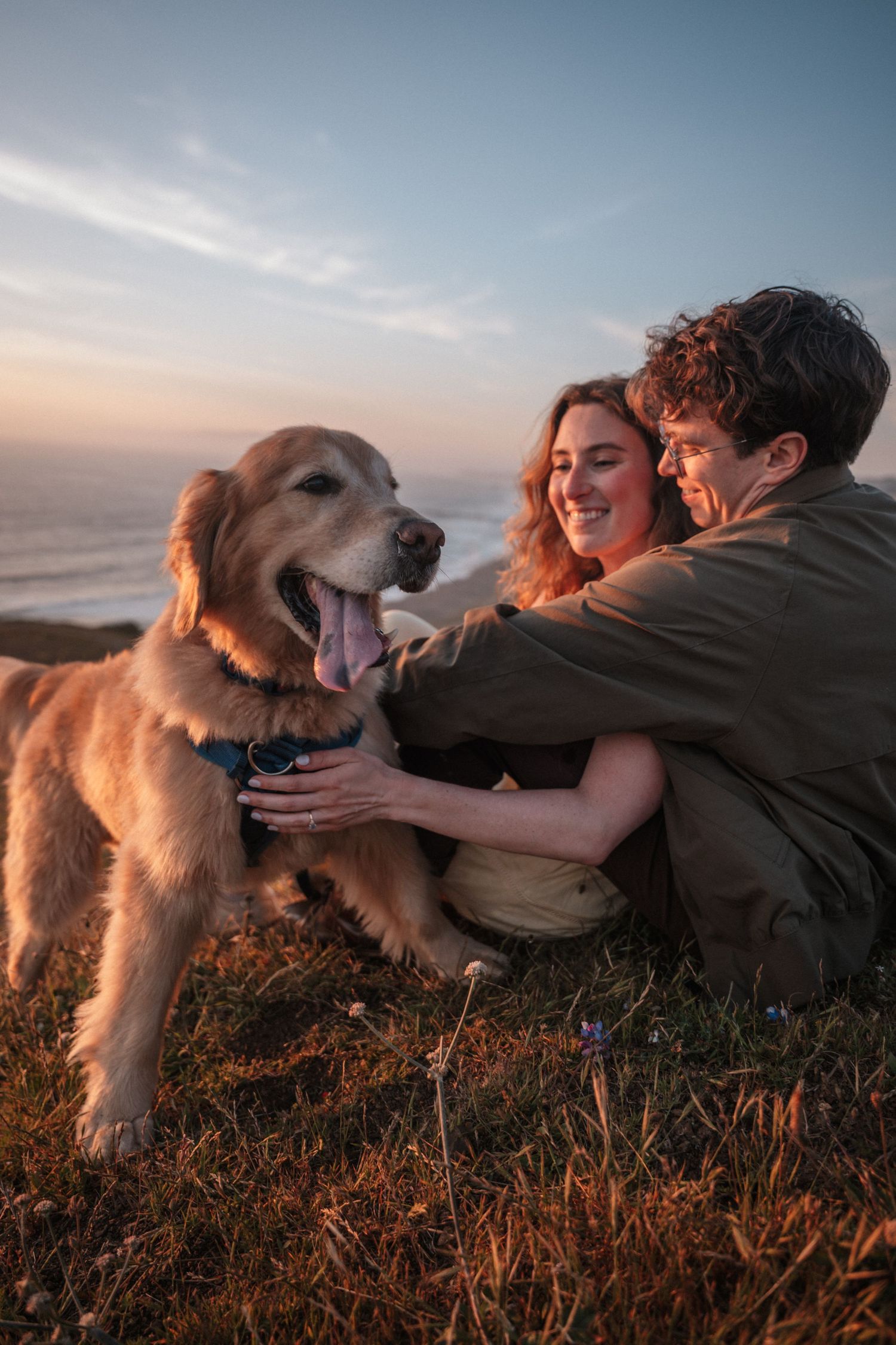 Happy couple sitting on a grassy cliff petting their golden retriever at dusk.