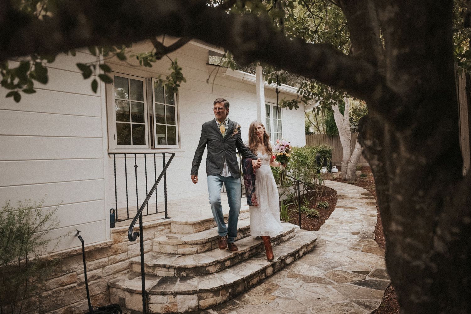 Couple in bohemian wedding attire walks down stone steps outside a white cottage surrounded by lush greenery.
