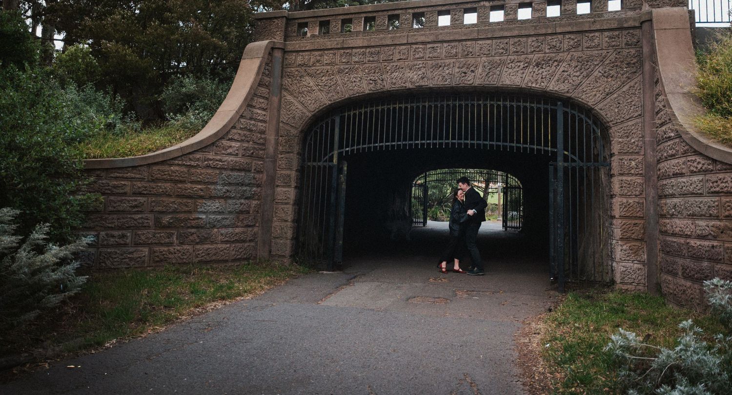Couple embracing under a stone arch bridge with iron gates in a lush park setting.