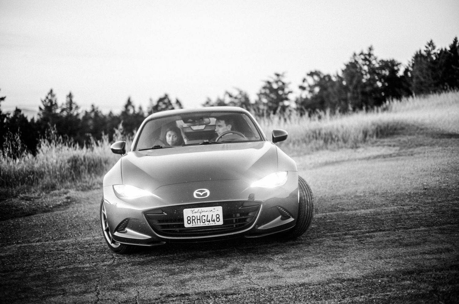 Black and white photo of a Mazda MX-5 Miata parked on a dirt road surrounded by trees.