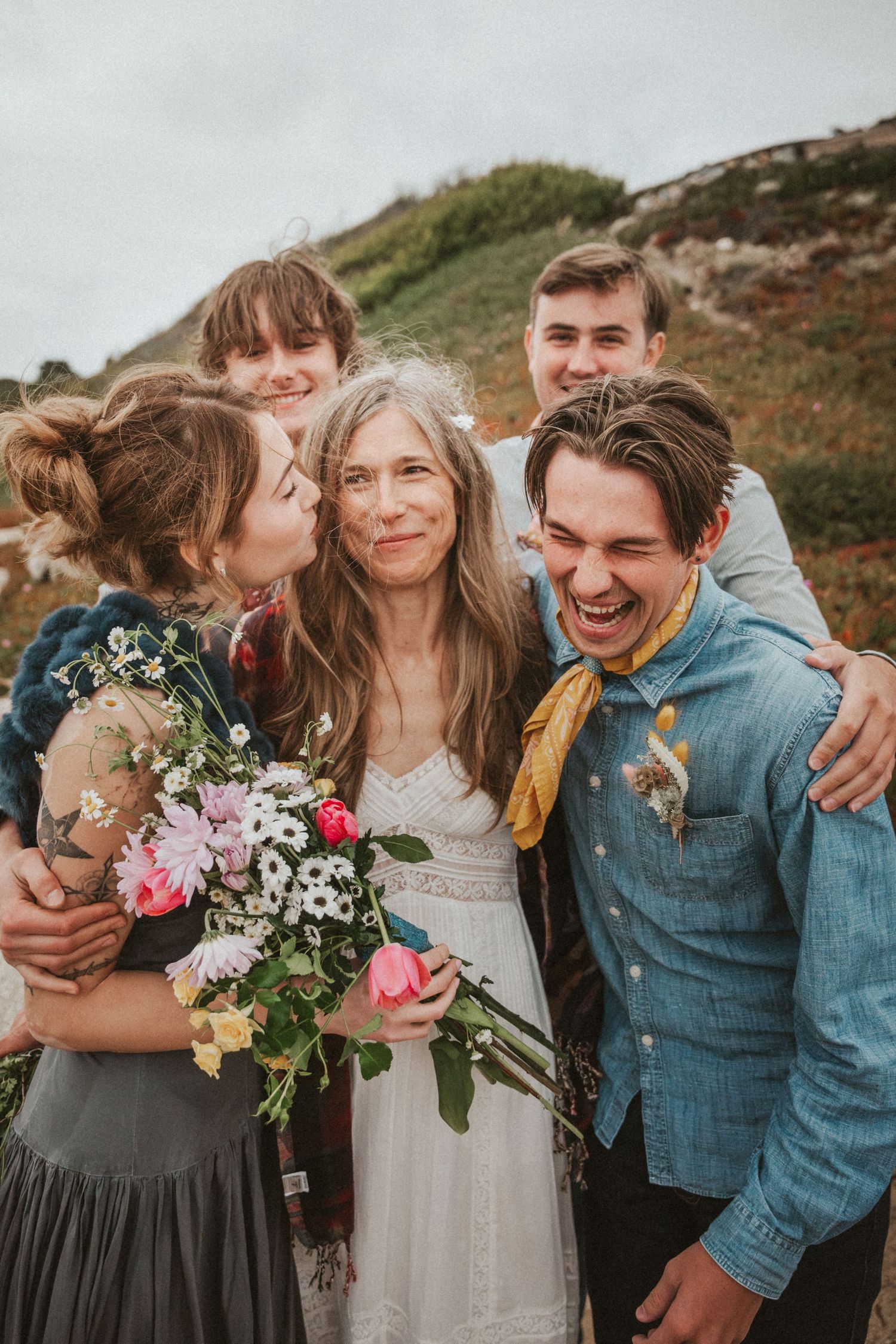 A bride in white dress surrounded by joyful family members outdoors on a hillside.