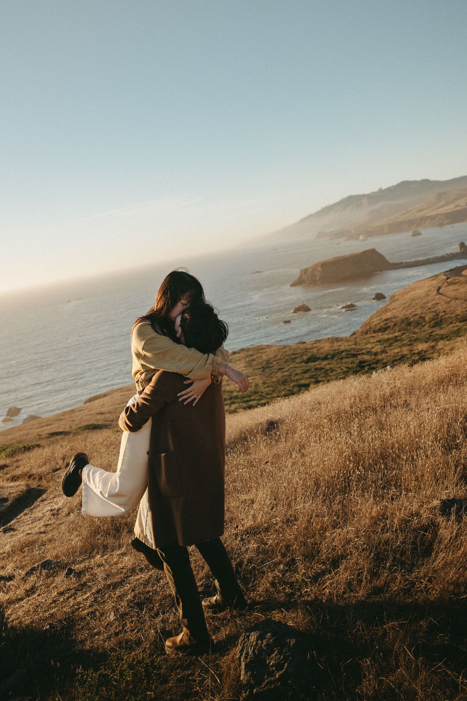 Couple embracing on a grassy coastal hillside at golden hour, one partner lifted off the ground, with the ocean and cliffs behind.