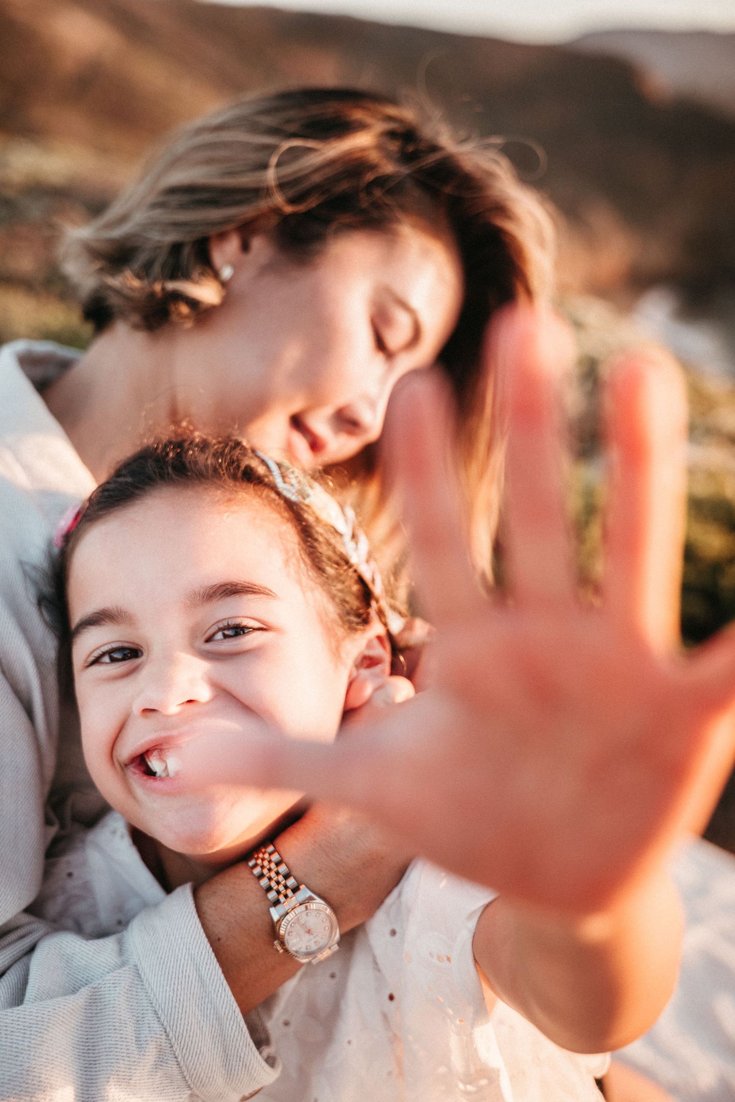 A mother tenderly holds her laughing daughter who reaches toward the camera outdoors.