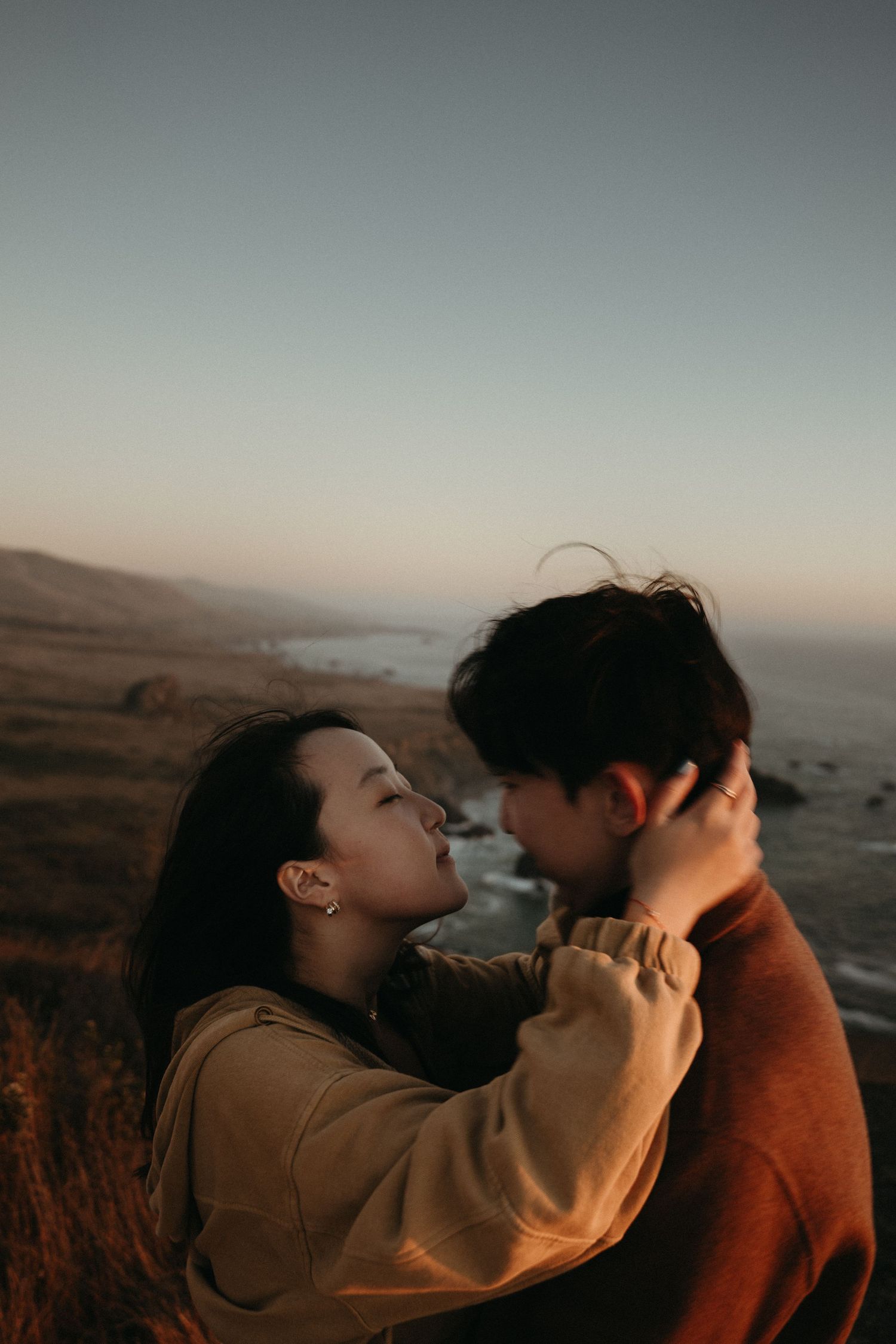 A couple embraces tenderly on a coastal bluff at sunset, with soft golden light and the ocean stretching behind them.