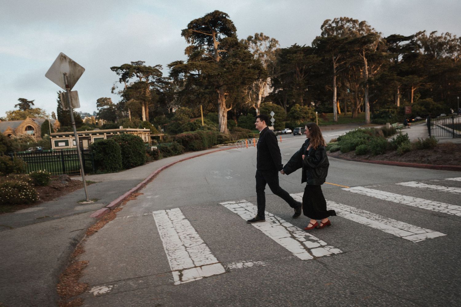 Couple holding hands crossing a zebra crosswalk on a quiet road surrounded by lush green trees.