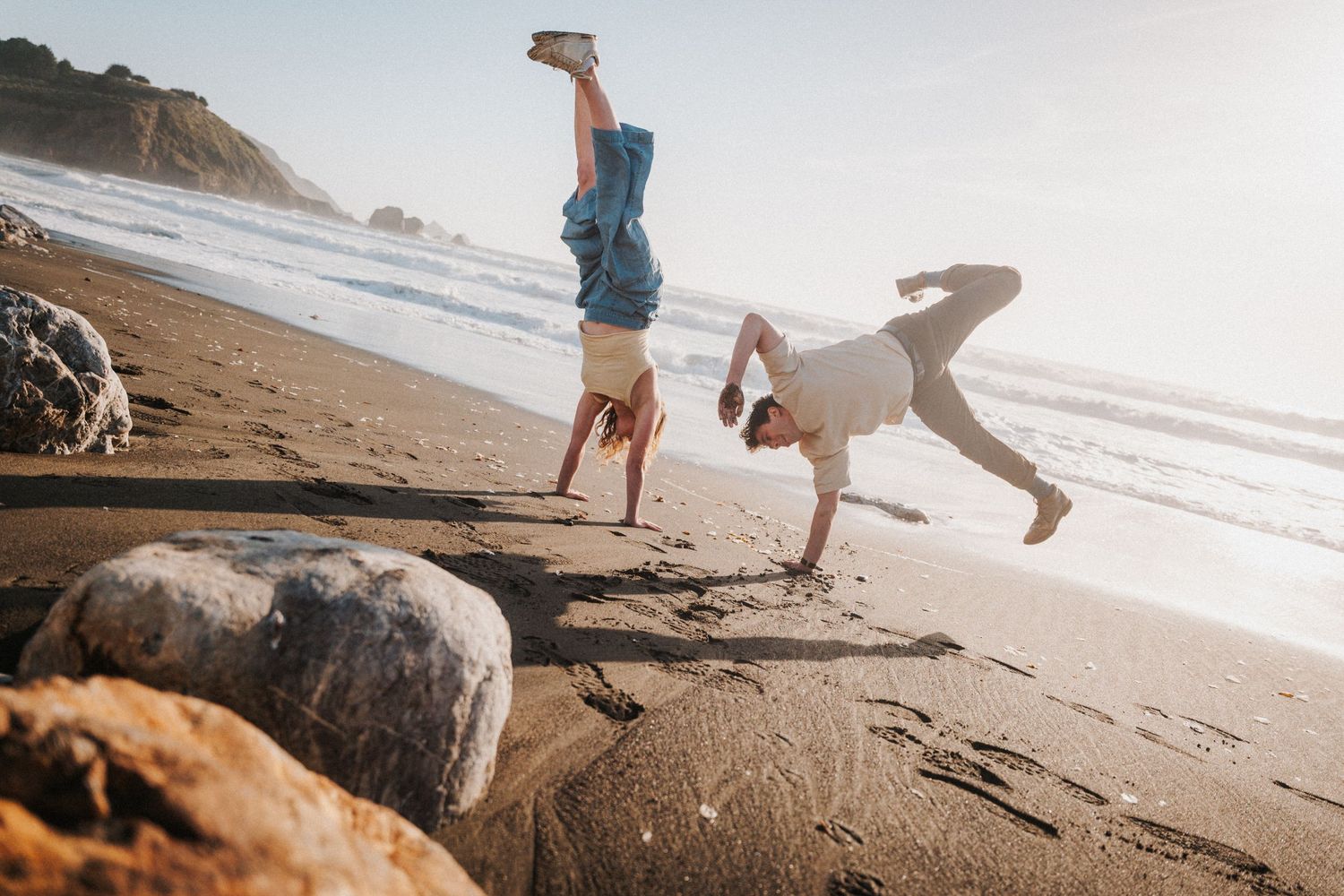 Two people doing handstands on a sandy beach with ocean waves and rocky coastline in golden sunlight.