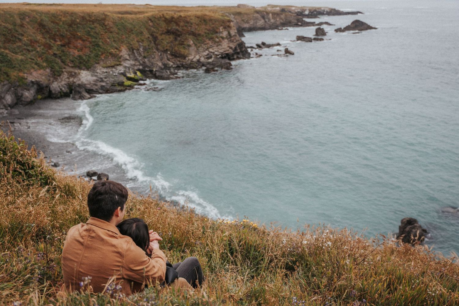 Couple sitting on a grassy coastal cliff overlooking a rocky shoreline and turquoise ocean waters.