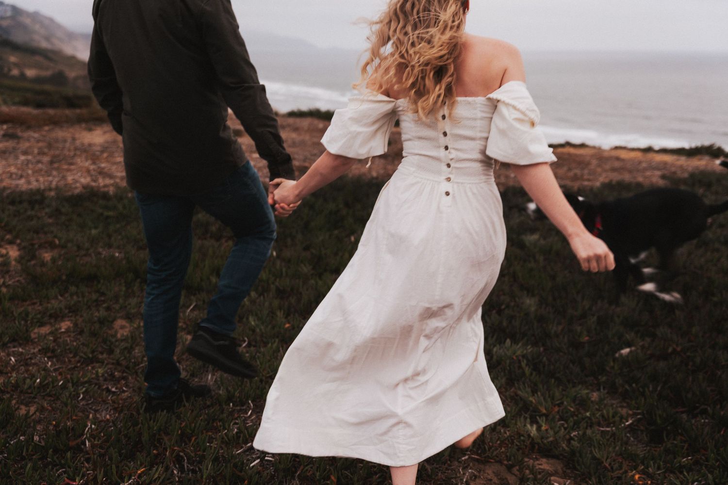 Couple holding hands running through coastal cliffside meadow, woman in flowing white off-shoulder dress with button back detail.