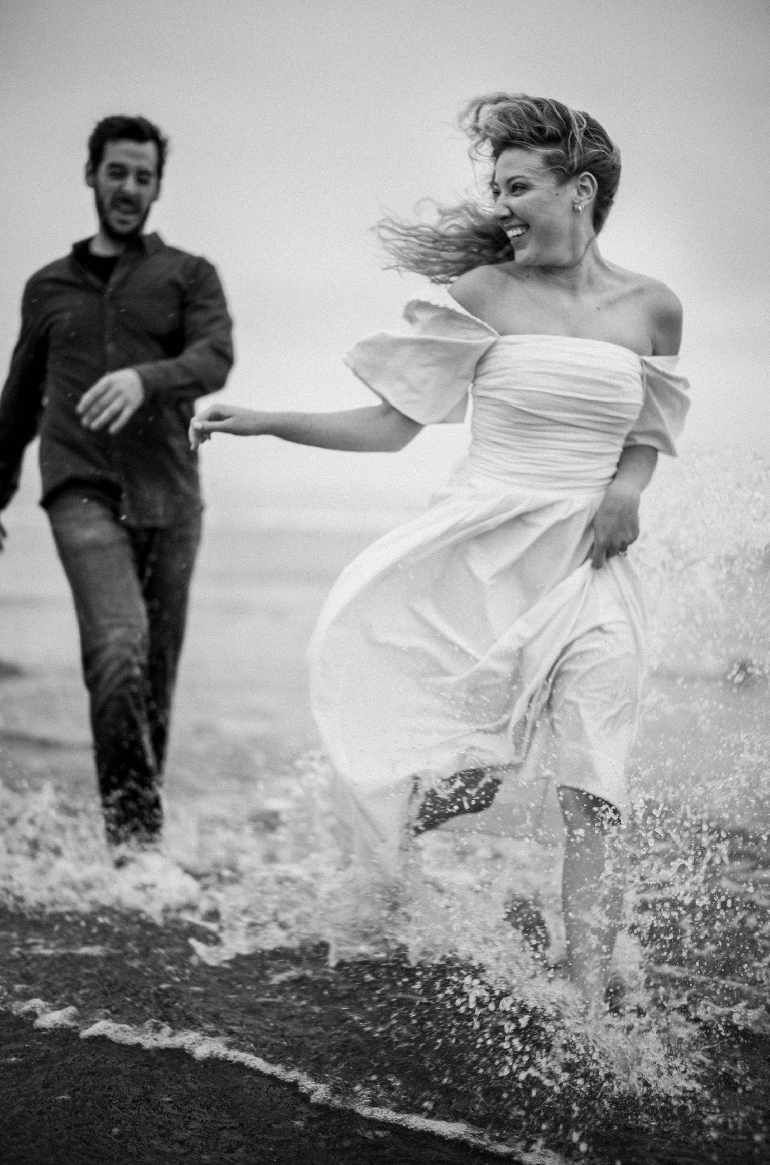 Black and white photo of a joyful bride in white dress splashing through ocean waves with groom.
