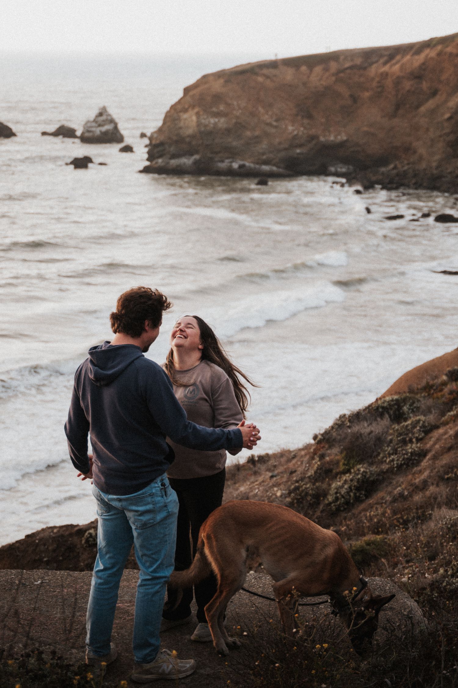 Happy couple holding hands and laughing by the ocean with their dog on a rocky cliff.