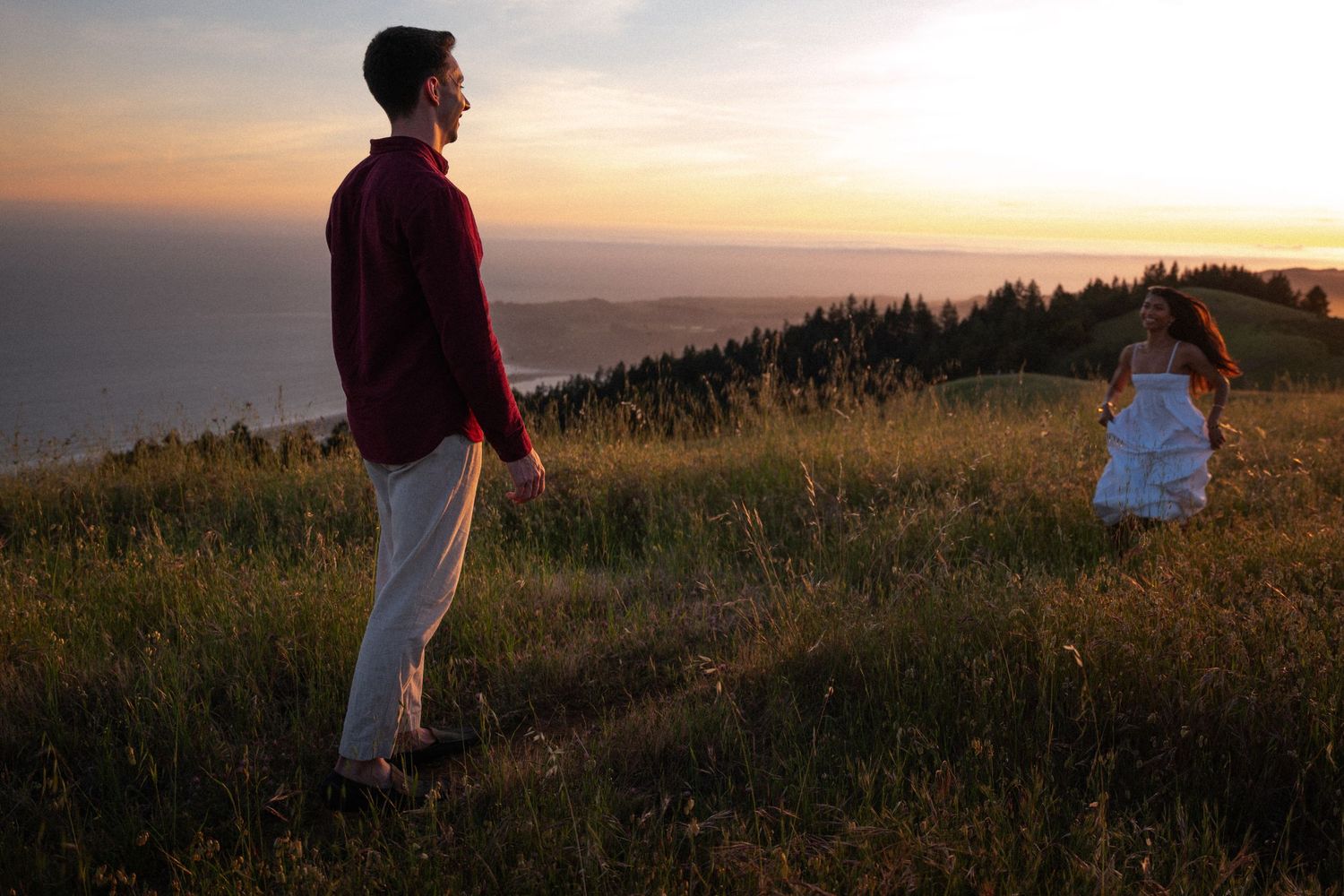 A man in a red shirt watches a woman in a white dress on a grassy hill at sunset with ocean views.