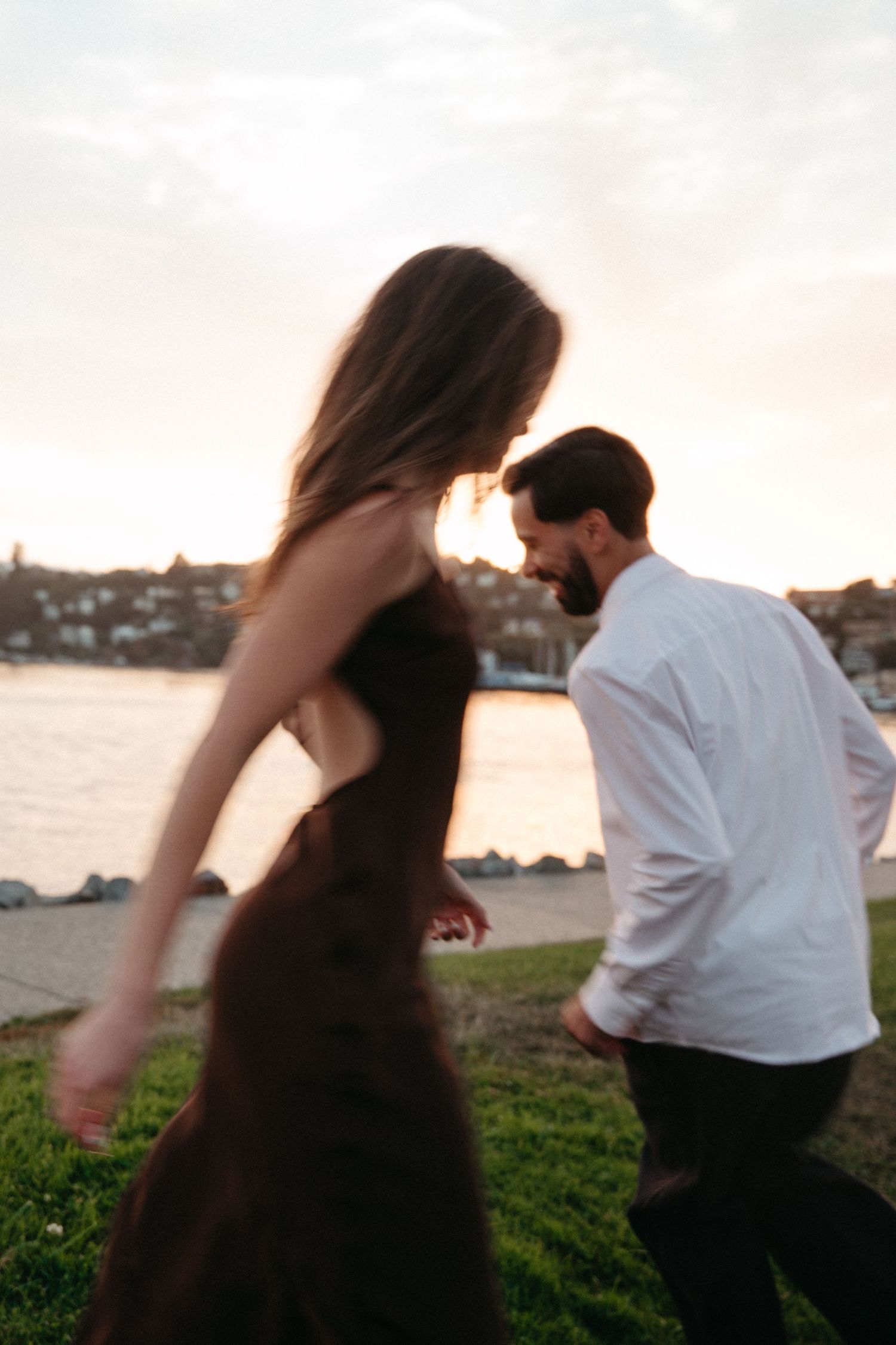 A couple dances romantically by a riverside at sunset, woman in brown dress and man in white shirt.