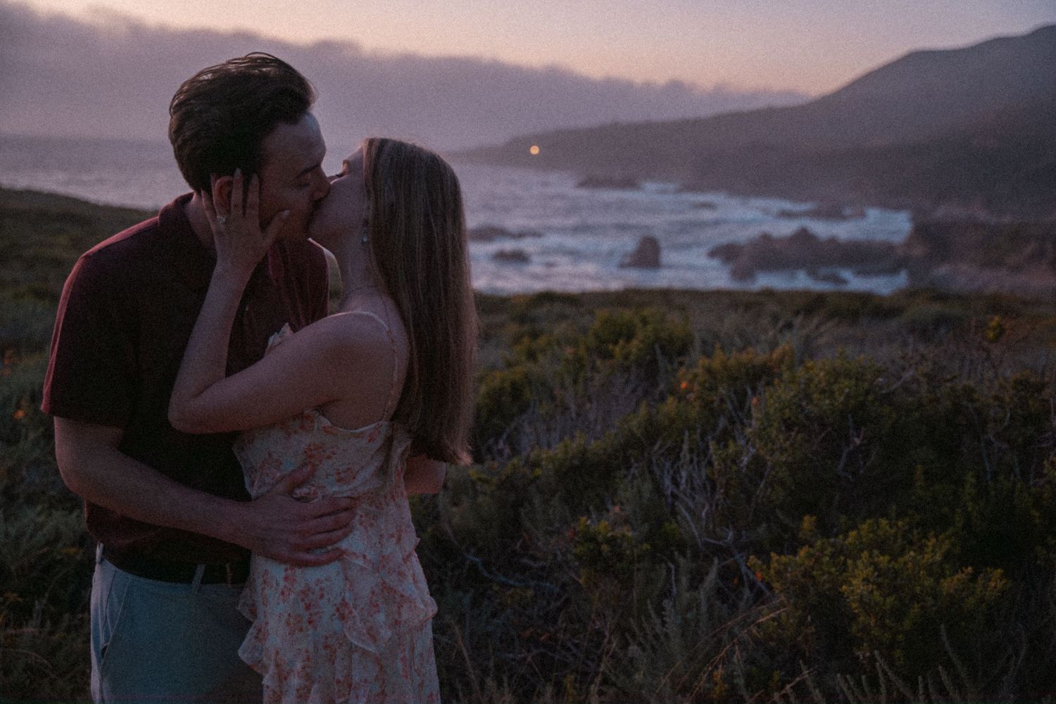 Couple sharing a romantic kiss at sunset on a scenic coastal overlook with ocean waves and misty mountains in the background.