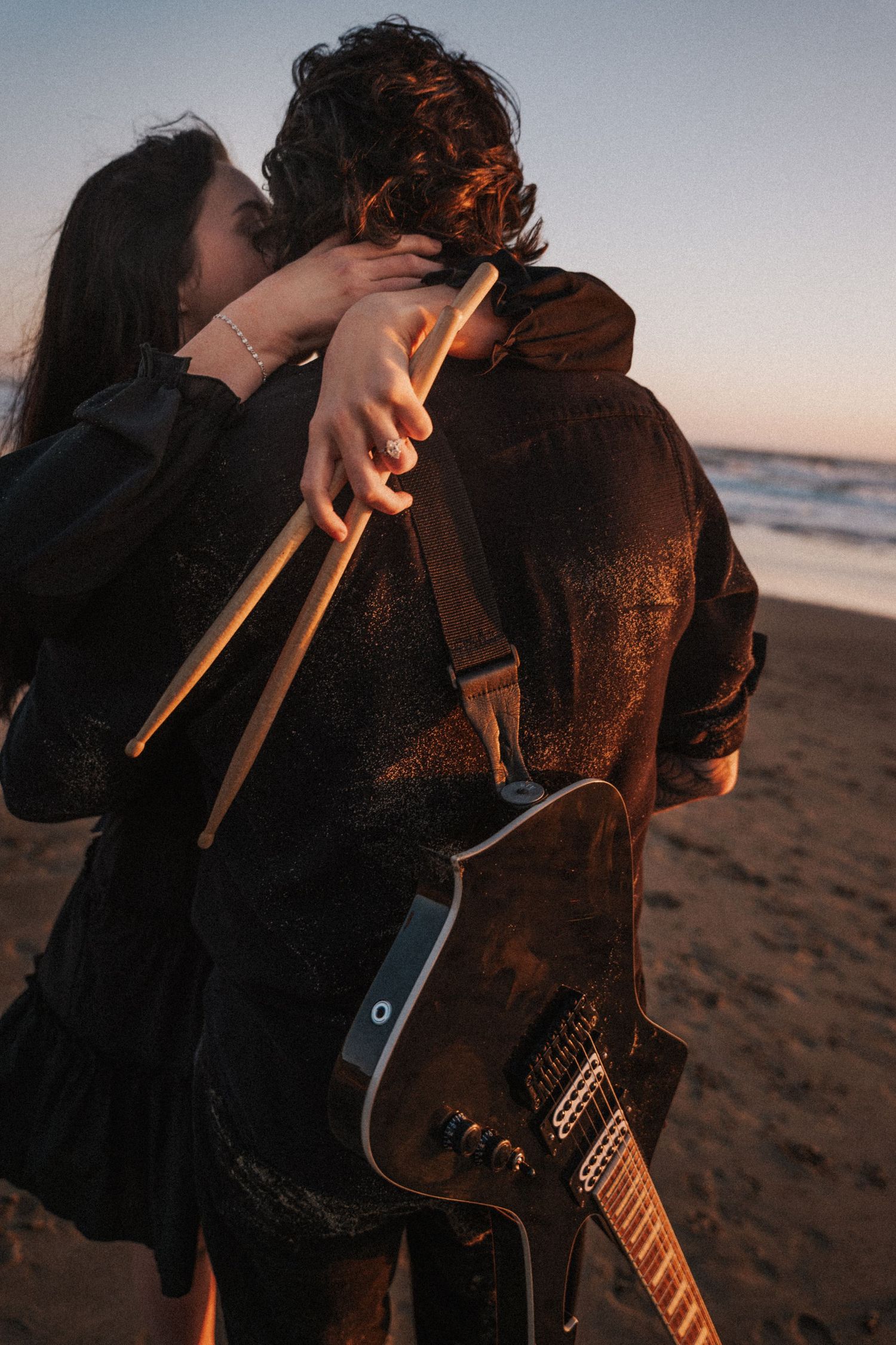 Couple embracing on beach at sunset, woman holding drumsticks, man carrying electric guitar on strap. Photography by The Y.K. Studio.