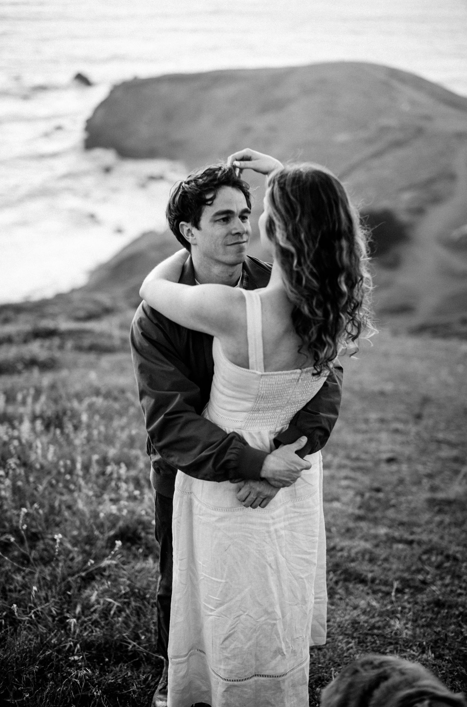 Black and white portrait of couple dancing intimately on coastal bluff with ocean and rocks behind them.