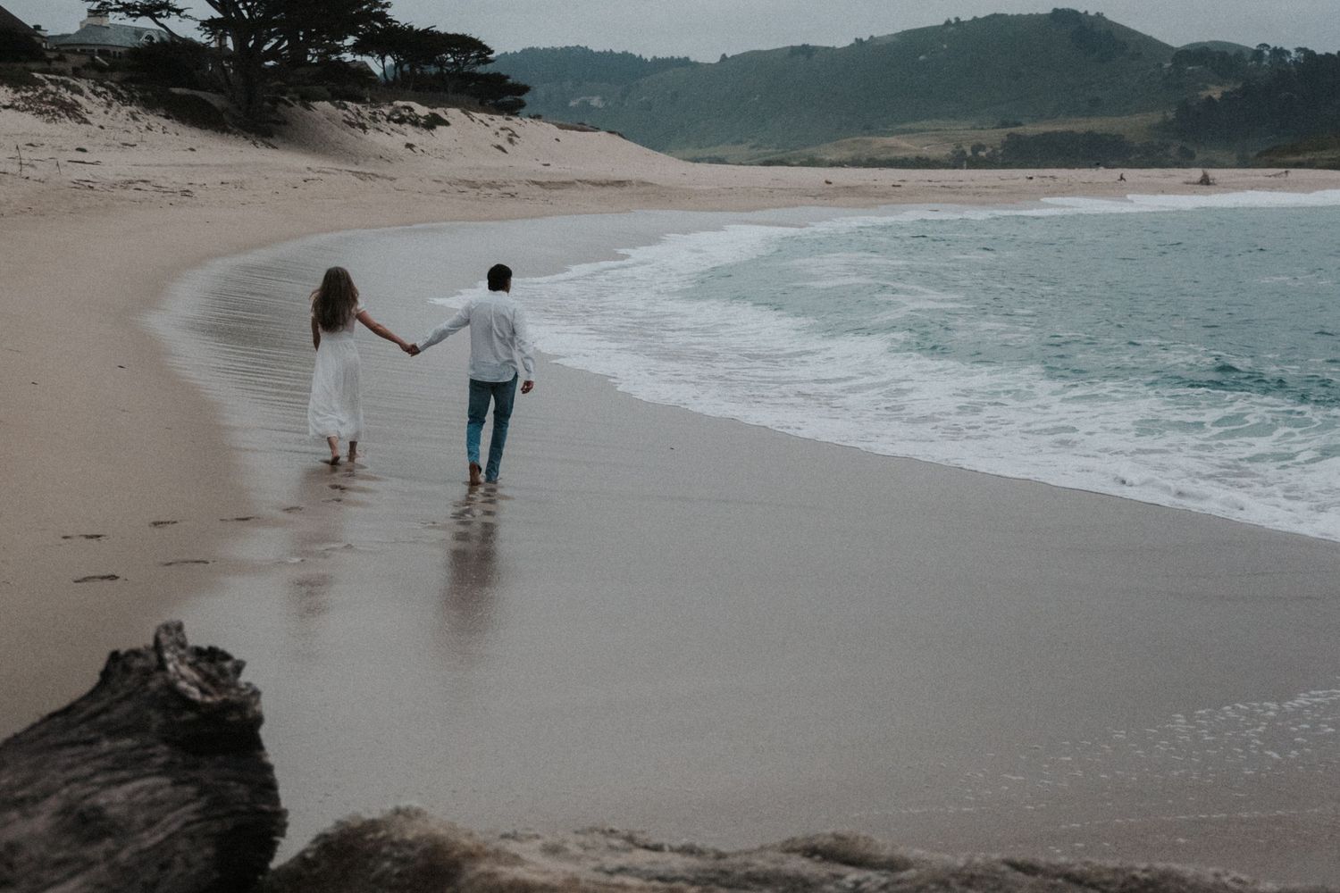 Couple holding hands walking along a sandy beach with turquoise waves, wearing white dress and jeans.