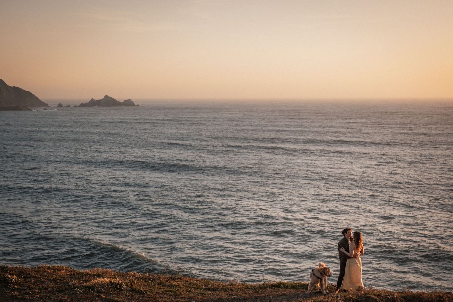 Couple sharing a romantic kiss at sunset on a coastal cliff overlooking the ocean with a small dog nearby.