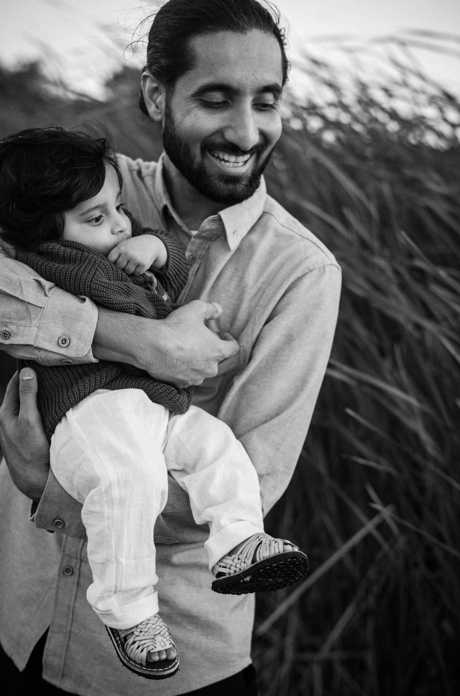 Black and white photo of a smiling bearded man holding a young child outdoors near tall grass.