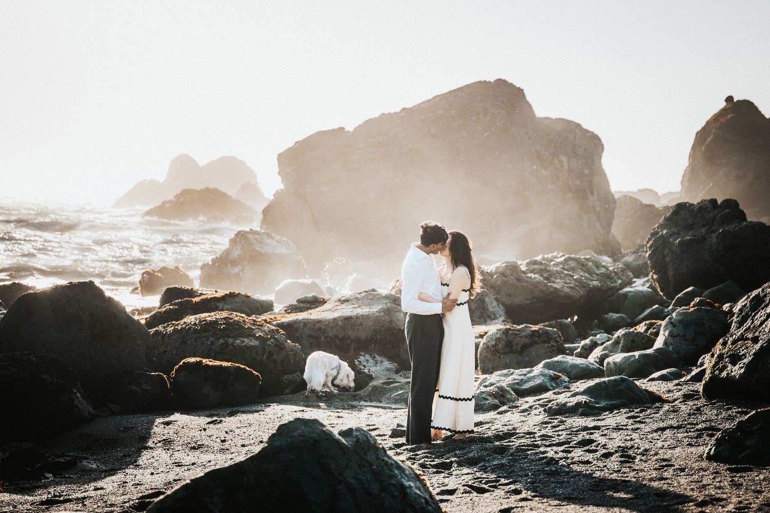 Couple embracing on rocky beach with misty ocean waves and large boulders in soft backlit sunlight.