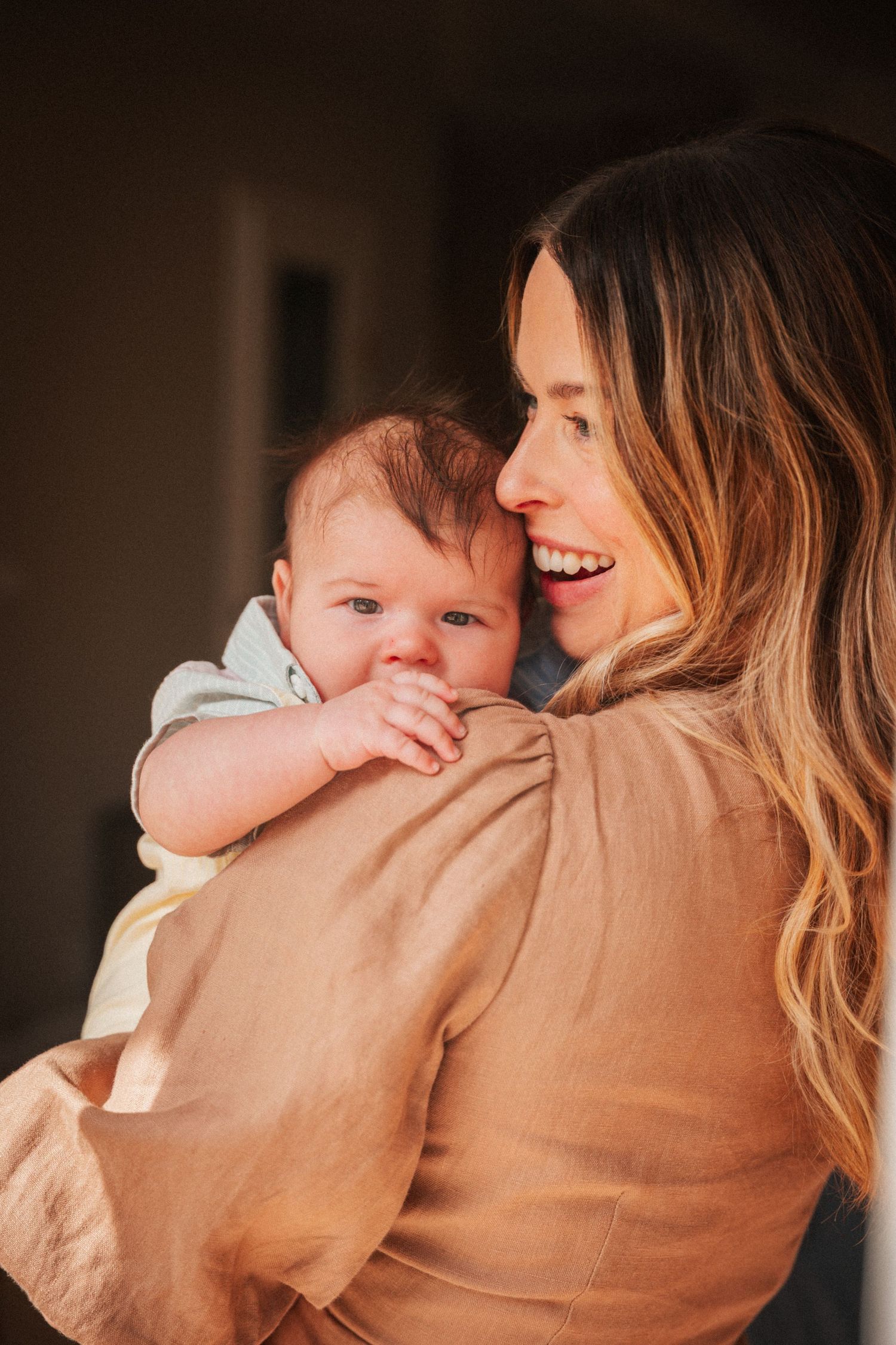 Smiling woman with long hair holding a cute baby in a sunlit room, sharing a tender moment.