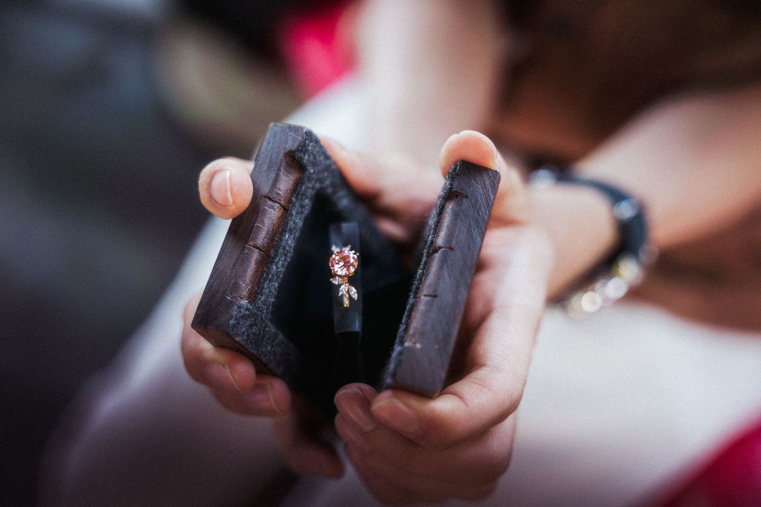 Hands holding open a dark wooden ring box displaying a delicate diamond engagement ring with pink gemstone accent.
