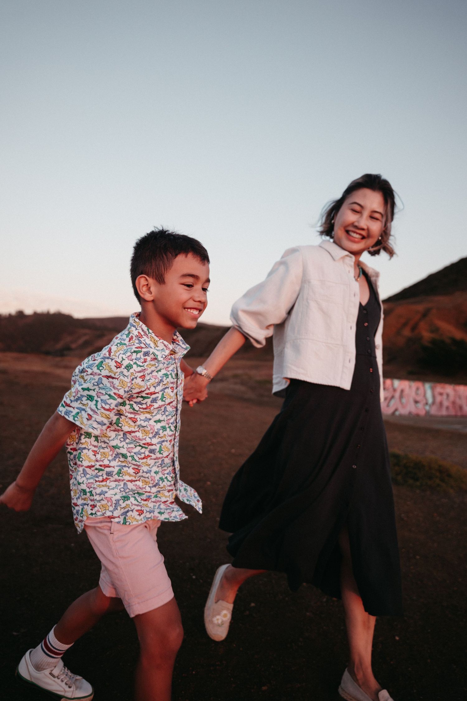 A joyful woman in a black dress and white jacket holds hands with a boy in a colorful shirt and pink shorts at sunset.