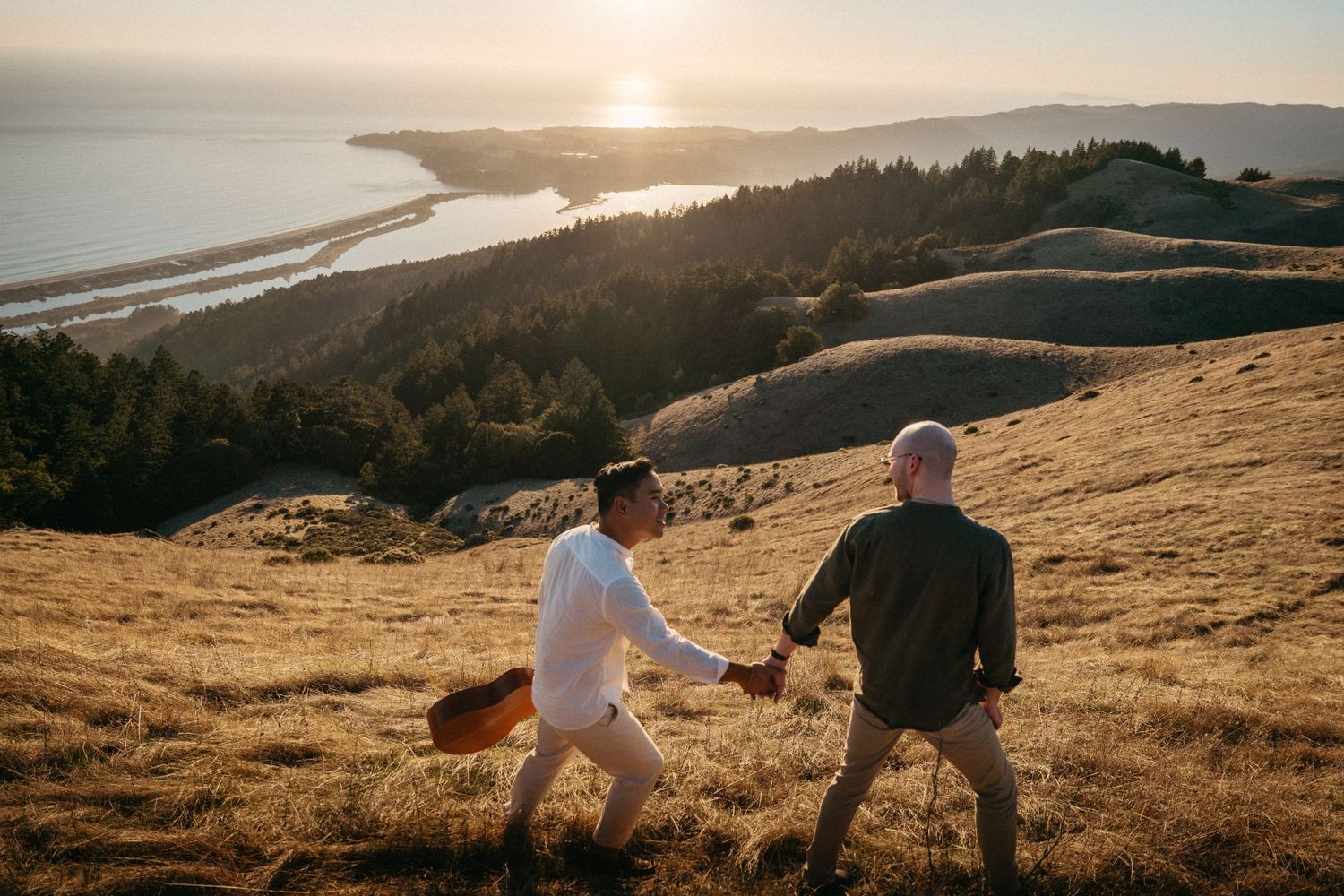 Two men hold hands walking up a golden hillside at sunset with a scenic coastal view in the background.