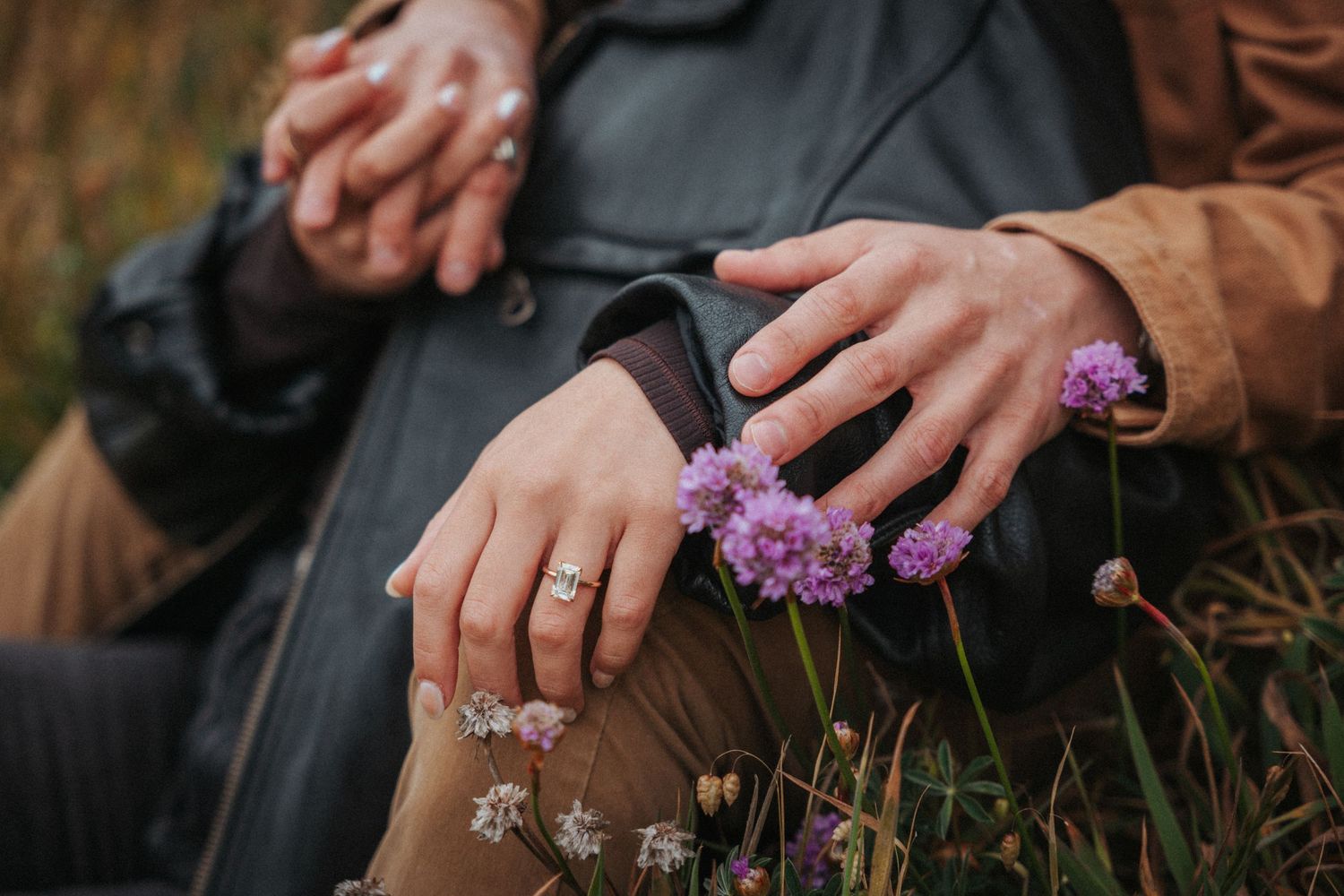 Close-up of couple holding hands with engagement ring and purple wildflowers in a field.