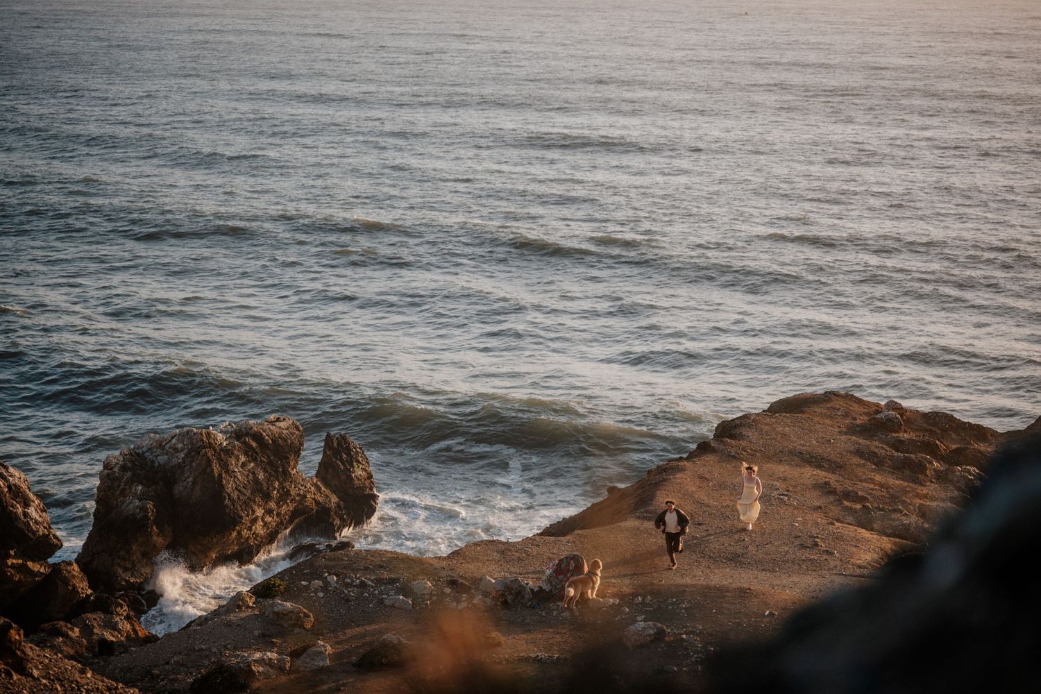 Couple in wedding attire standing on rocky coastal cliffs with ocean waves crashing below at golden hour.
