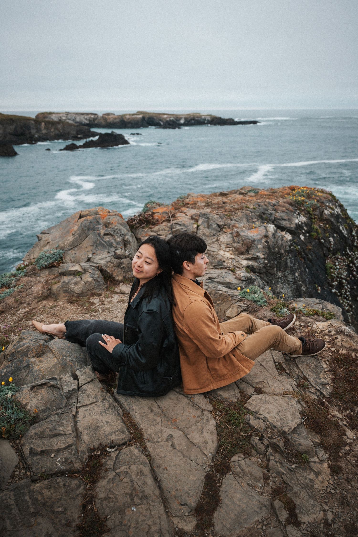 Two people sit back-to-back on coastal rocks overlooking a misty ocean, wearing casual earth-tone outfits.