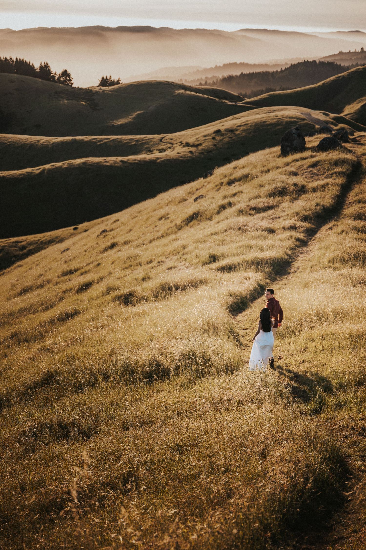 Couple walking through golden rolling hills at sunset in white dress and dark jacket.Photography by The Y.K. Studio.