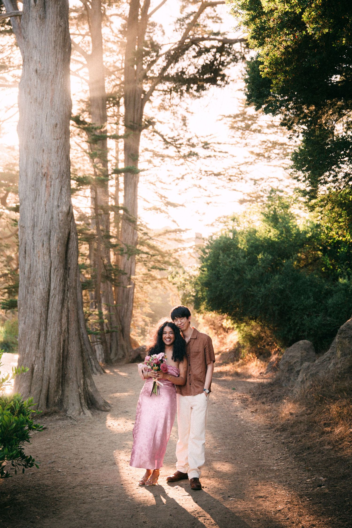 Couple embracing on a sunlit forest path surrounded by tall trees, woman in pink dress holding flowers.