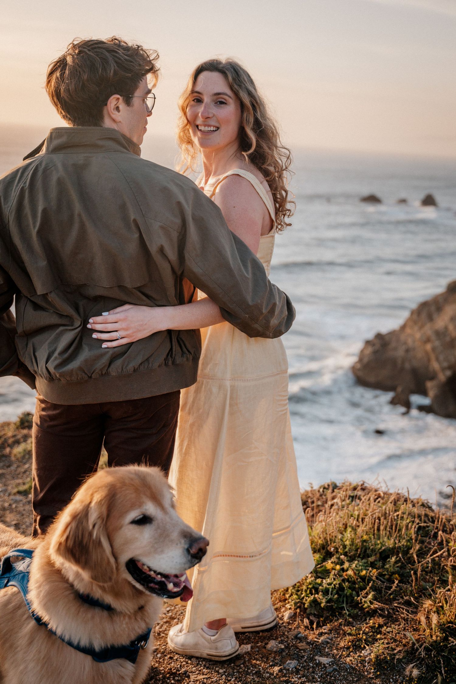 A couple embraces on a coastal cliff at sunset with their golden retriever dog nearby.