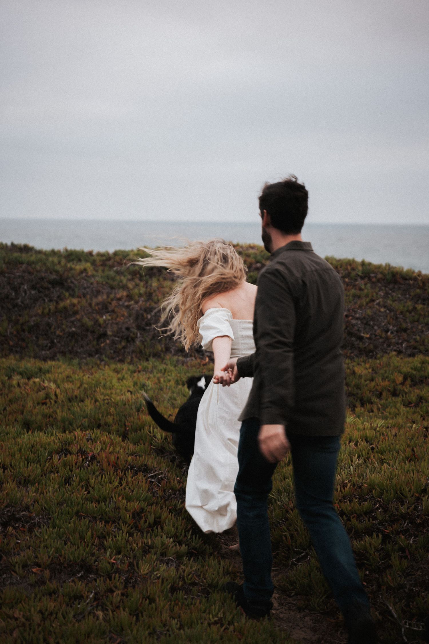Couple holding hands walking through coastal meadow, woman in white dress with windswept hair, overcast sky.