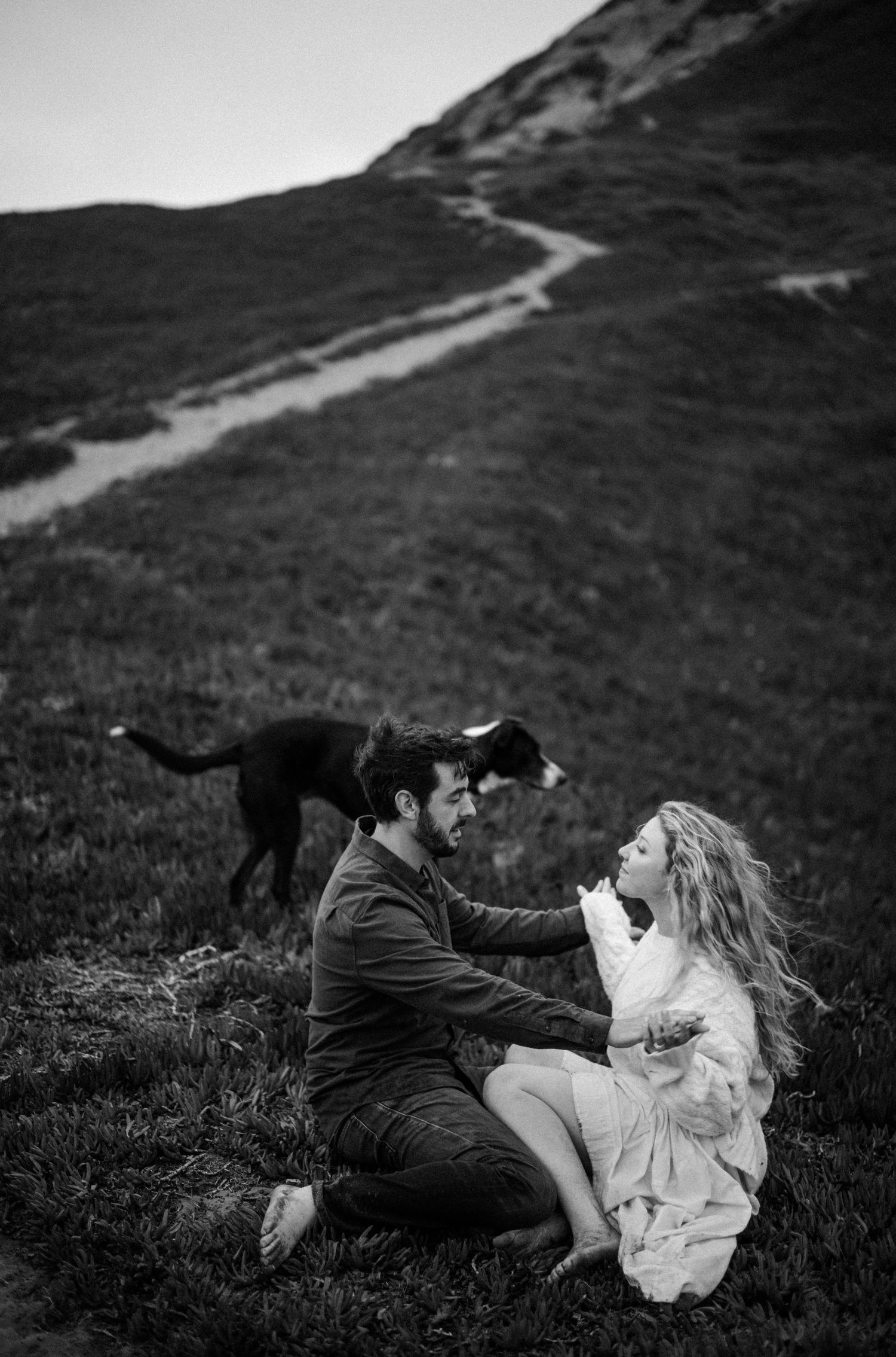 Black and white photo of a couple sitting on a grassy hillside with their dog in the background.
