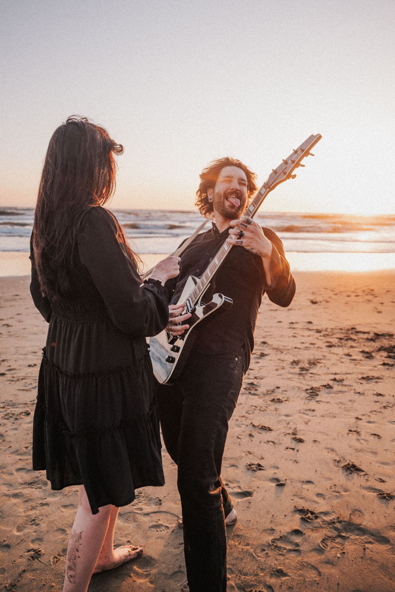 A smiling man plays electric guitar for a woman on a sandy beach at sunset, both dressed in black.