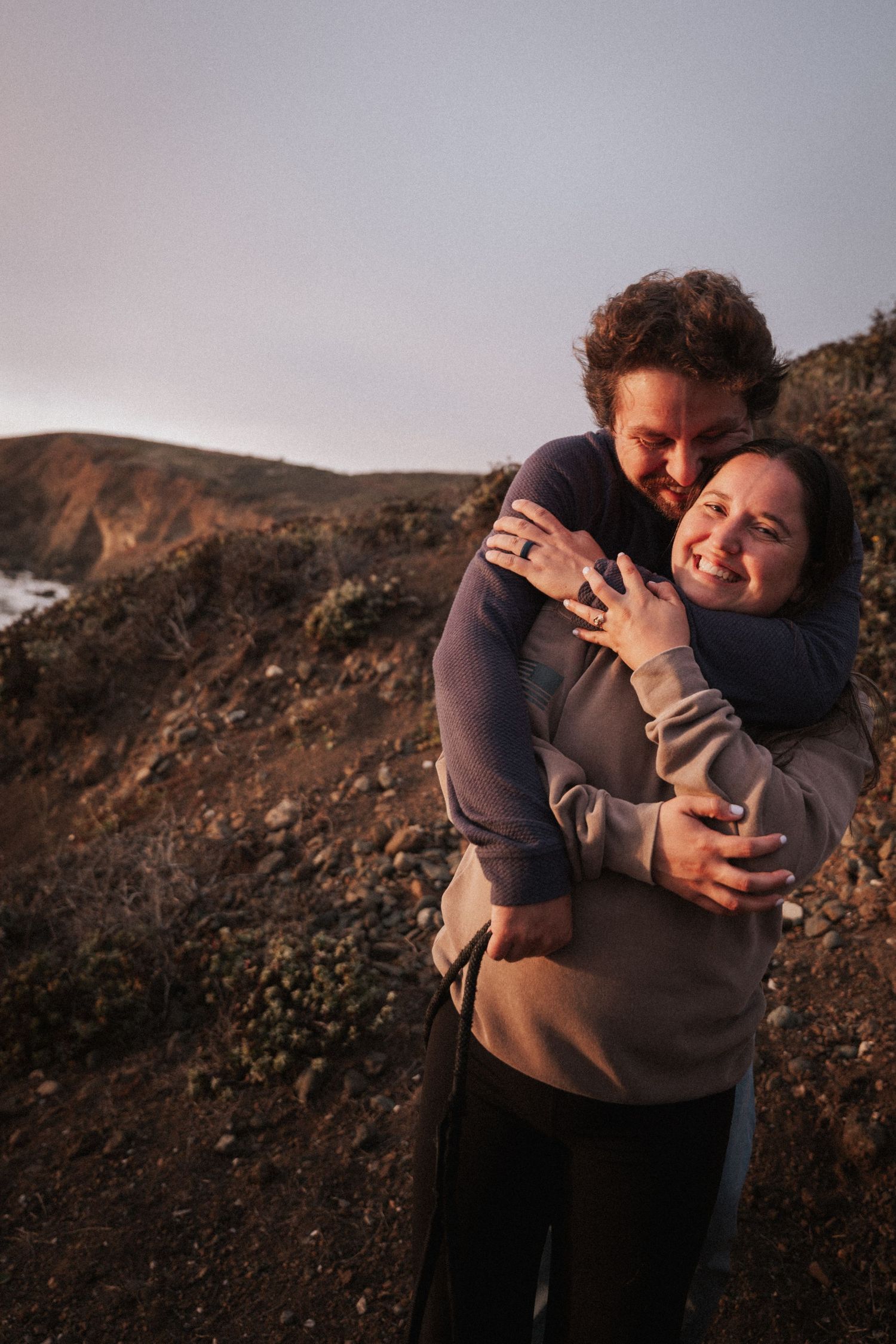 Couple embracing and smiling on a rocky hillside at golden hour, woman displaying an engagement ring.