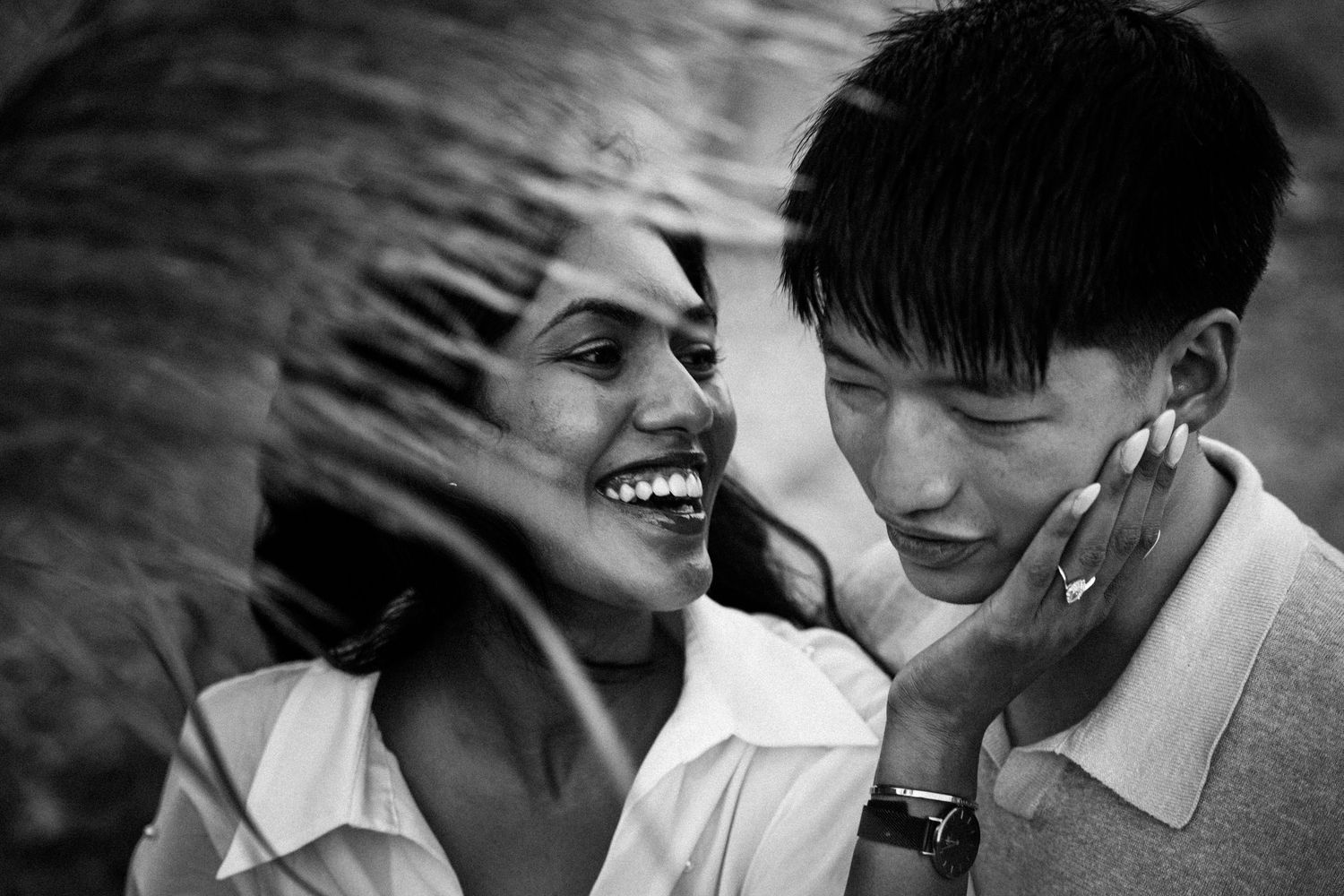 Black and white photo of a smiling couple sharing a joyful moment outdoors, with wind-blown hair adding movement.