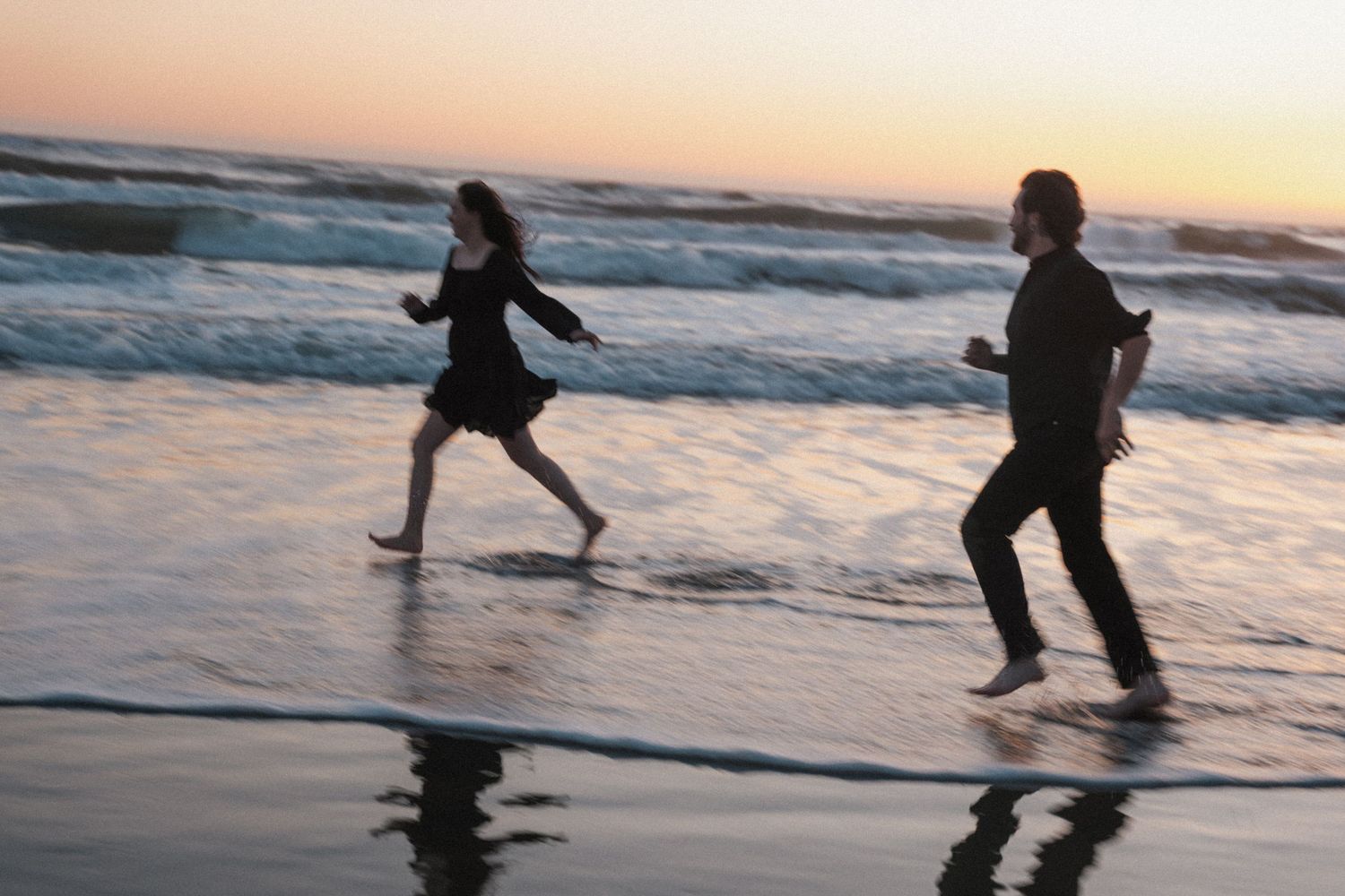 Two people running barefoot along a sandy beach at sunset with ocean waves in the background.