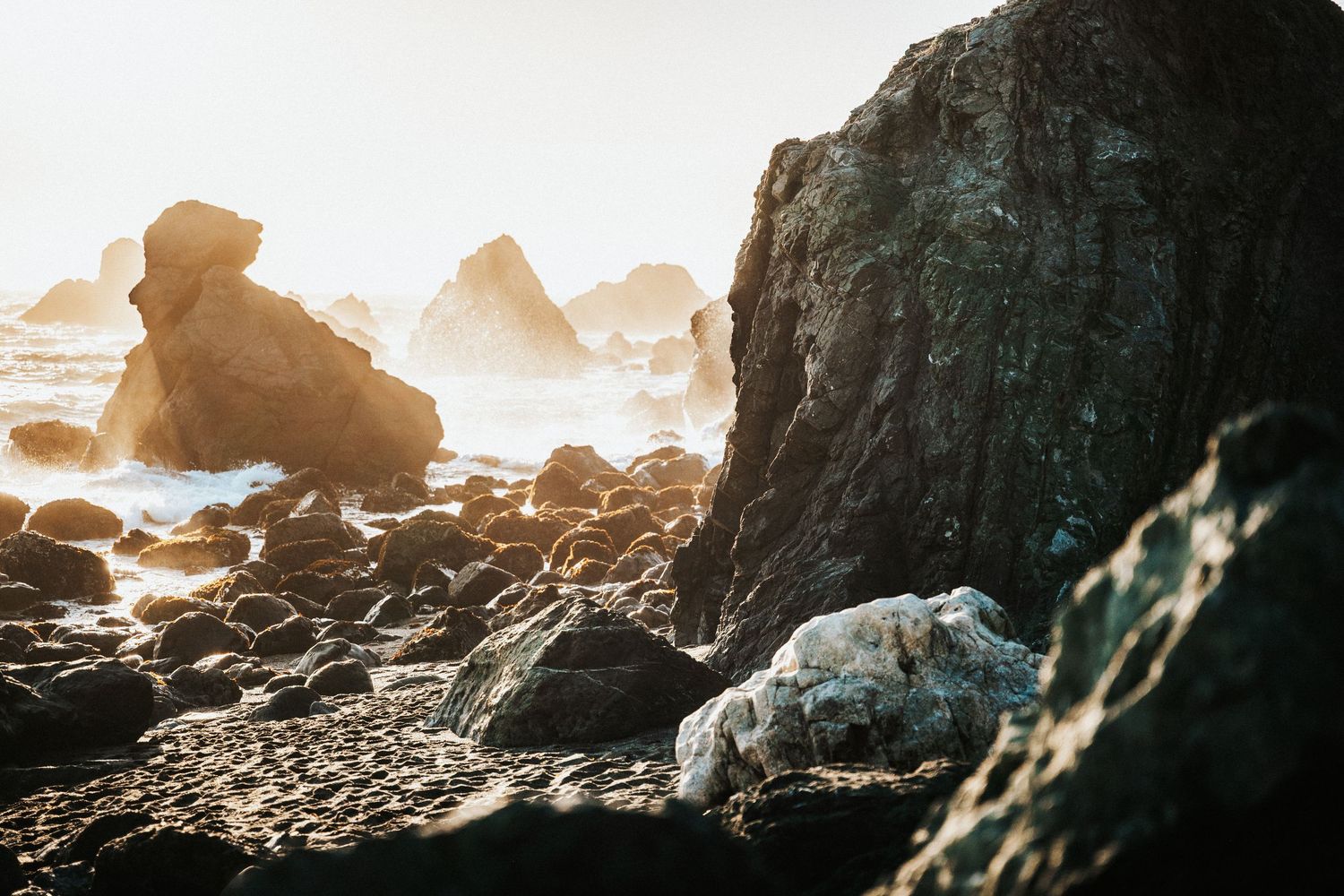 Rocky coastal shoreline with large boulders and misty ocean waves in warm golden sunlight.