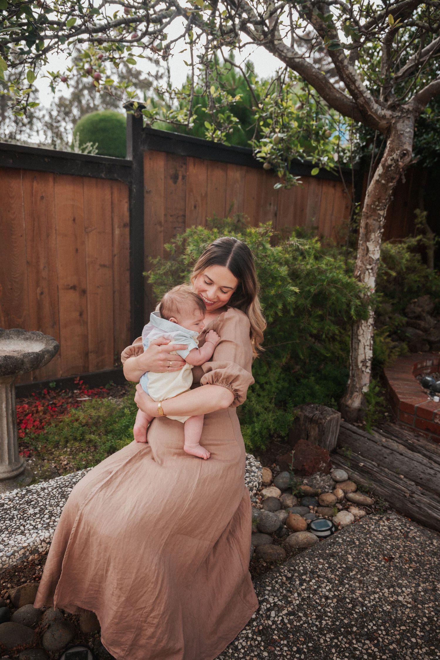 A mother in a blush maxi dress tenderly holds her newborn baby in a lush garden setting.