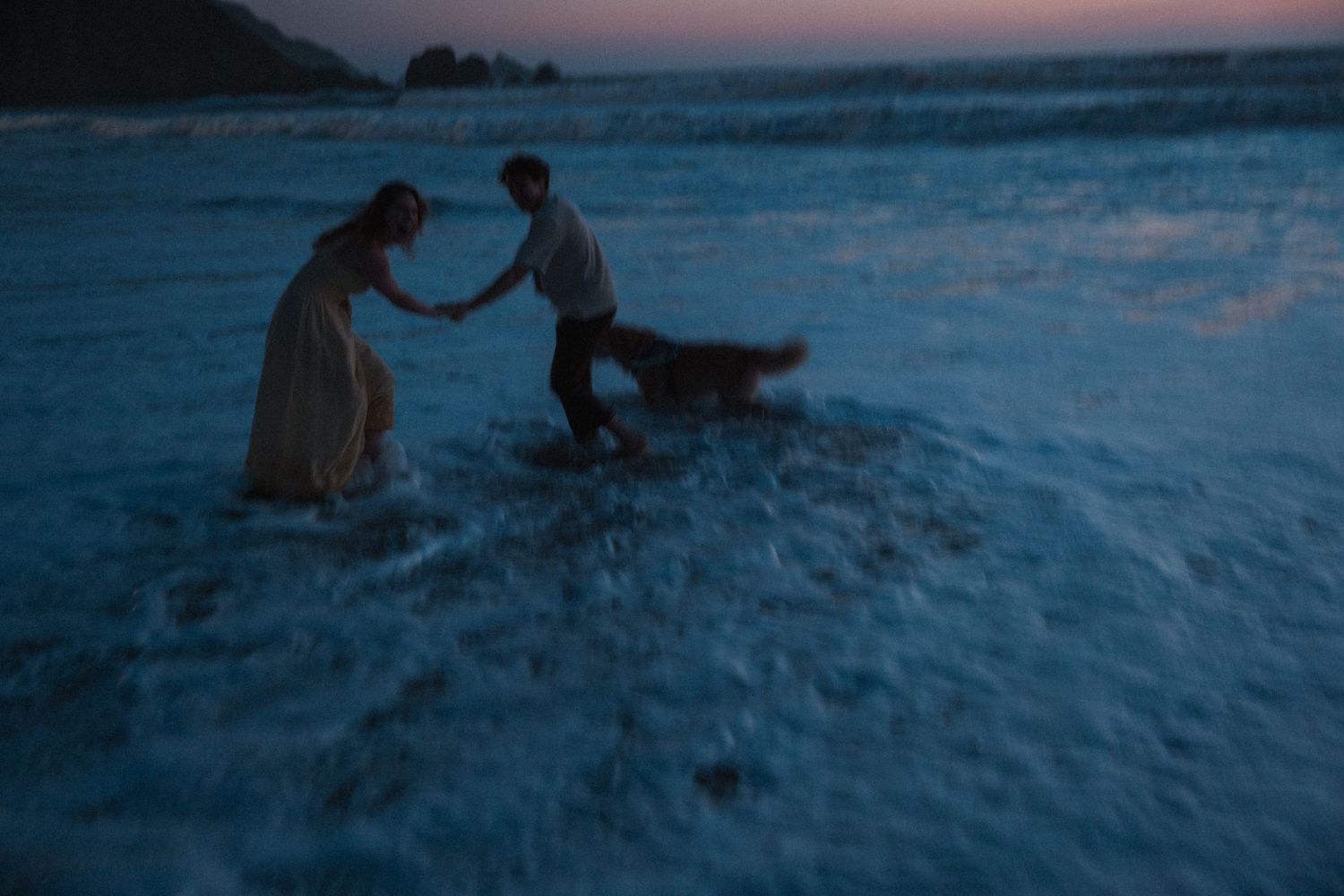 Couple and dog playing in ocean waves at dusk, blue twilight light on the beach.