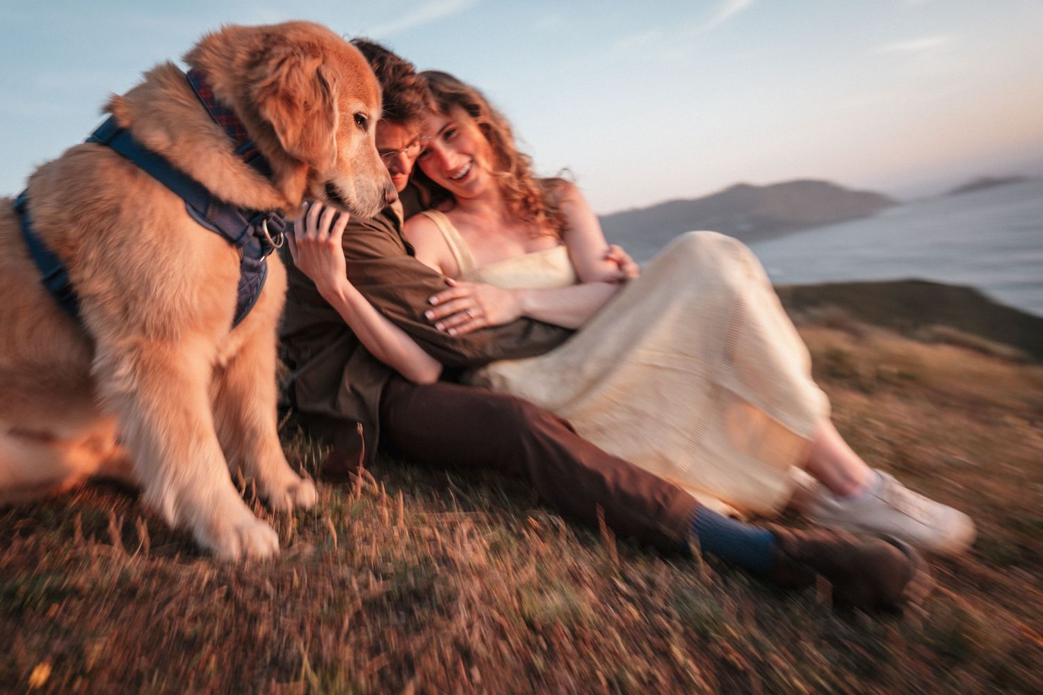 Couple sitting on a hillside laughing with a golden retriever dog, scenic coastal view at sunset. Photography by The Y.K. Studio.
