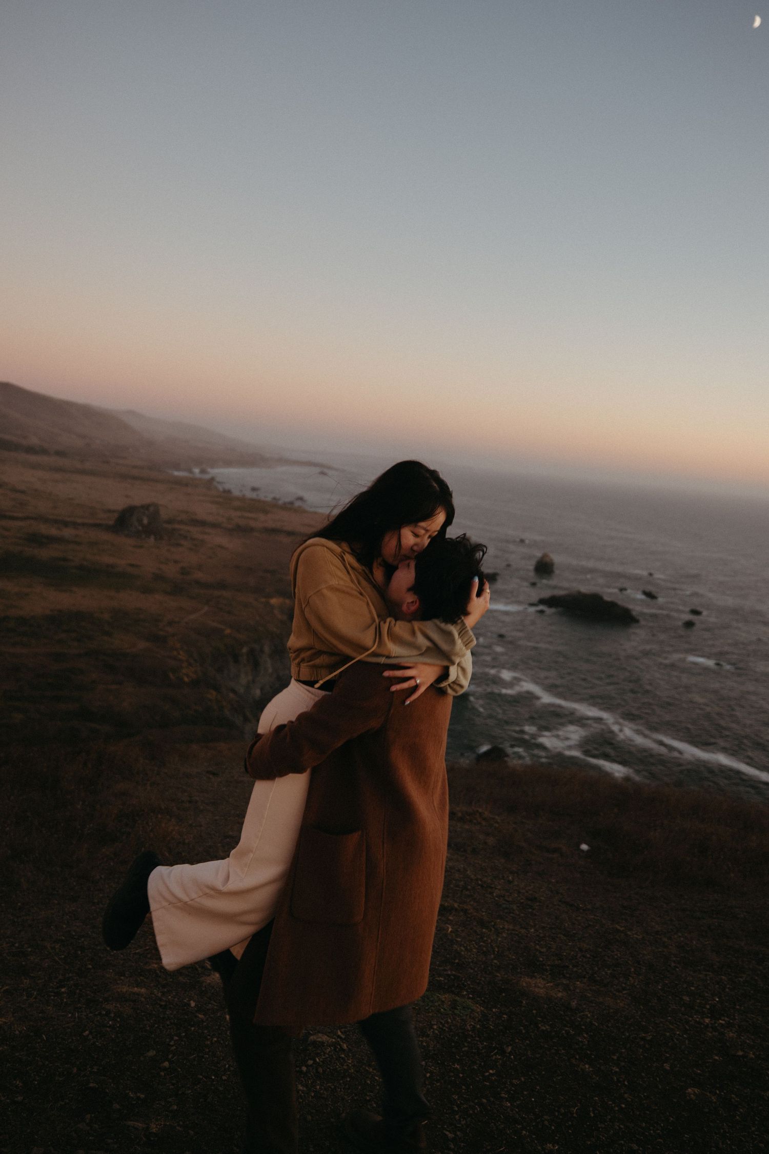 A woman in a mustard sweater wraps her arms around a man in a brown coat on a grassy coastal cliff at dusk.