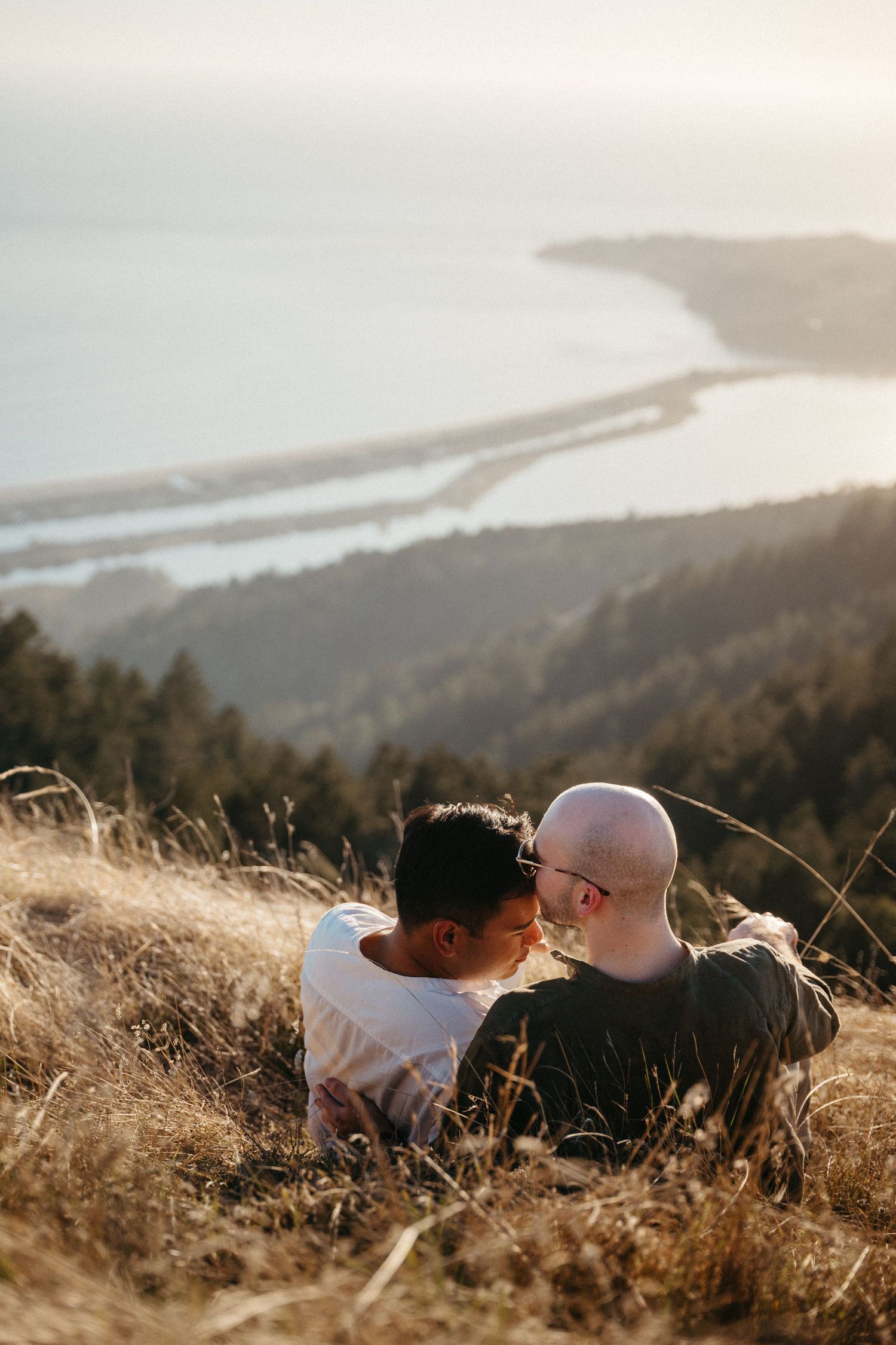 Two men sit closely together on a hillside overlooking a coastal landscape at golden hour.