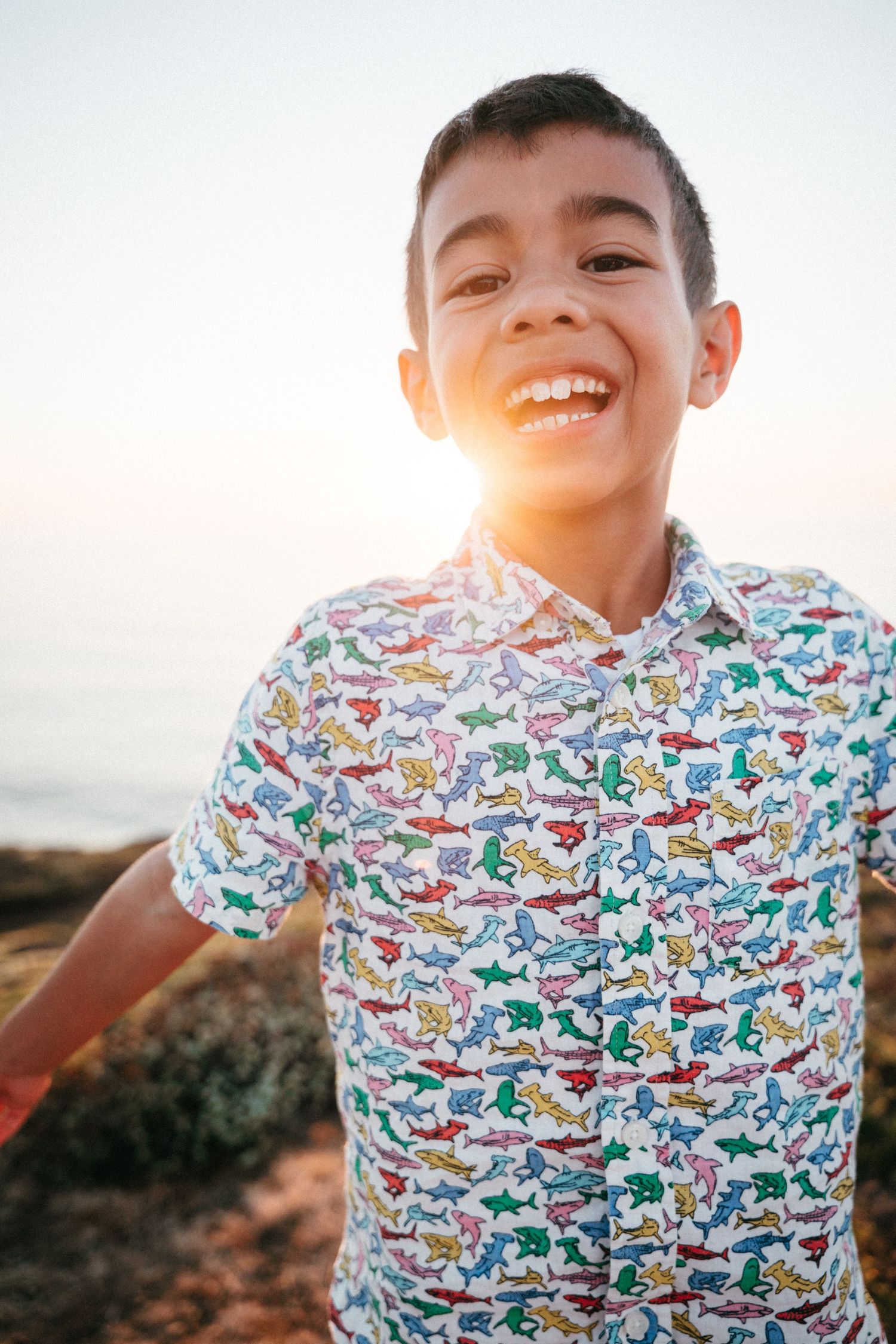 Smiling boy in colorful shark print shirt outdoors at sunset near the ocean.