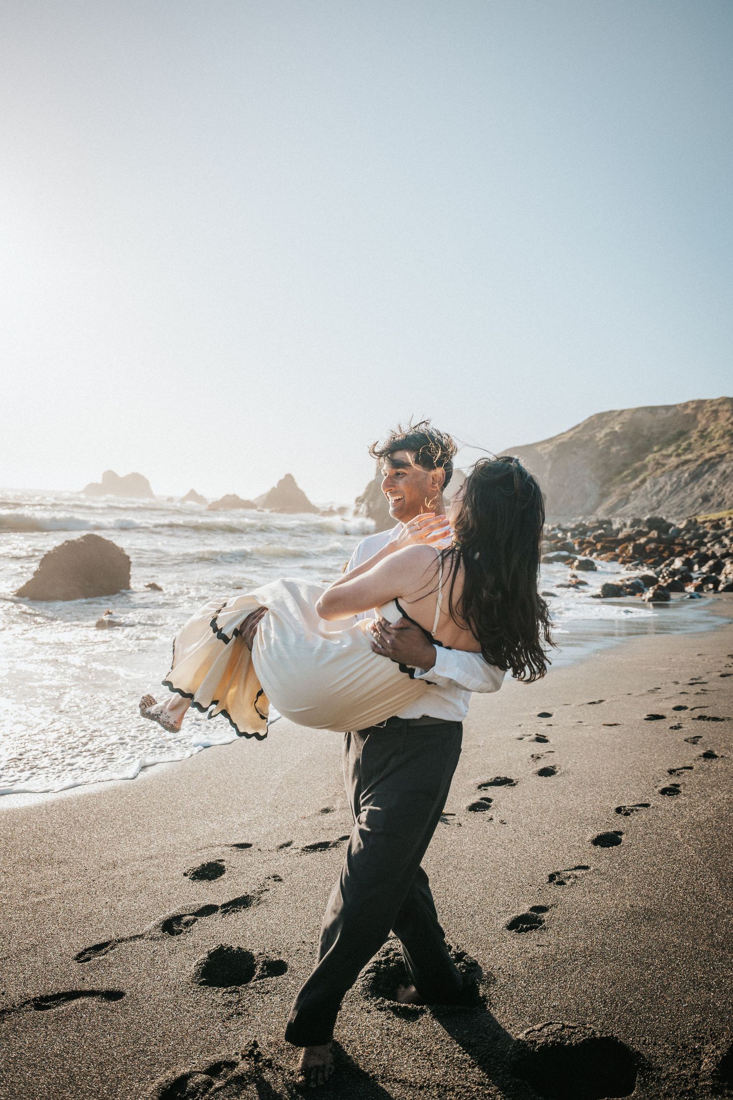 A groom carries his bride in his arms on a scenic beach at sunset, with ocean waves and rocky cliffs in the background.