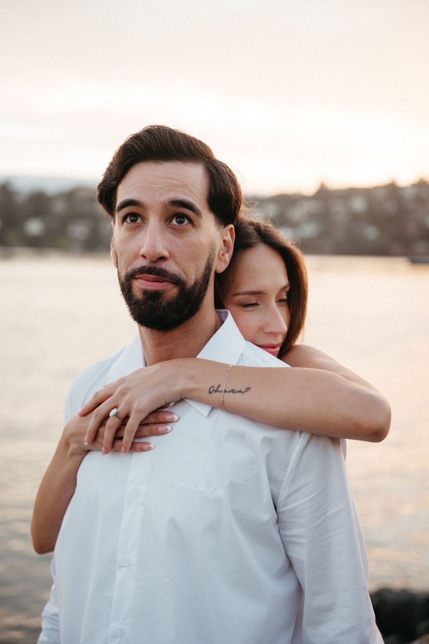 A bearded man in a white shirt is embraced from behind by a woman near a waterfront at sunset.